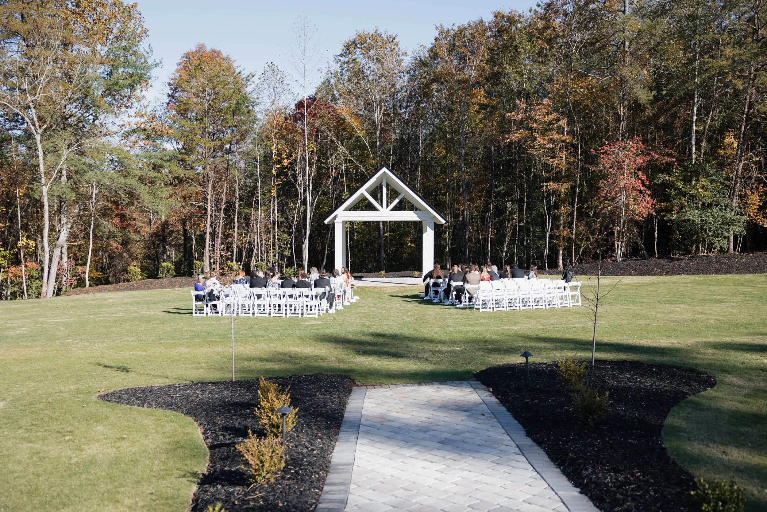 An outdoor wedding setup with white chairs facing a white arbor in a grassy area, surrounded by trees with autumn foliage.