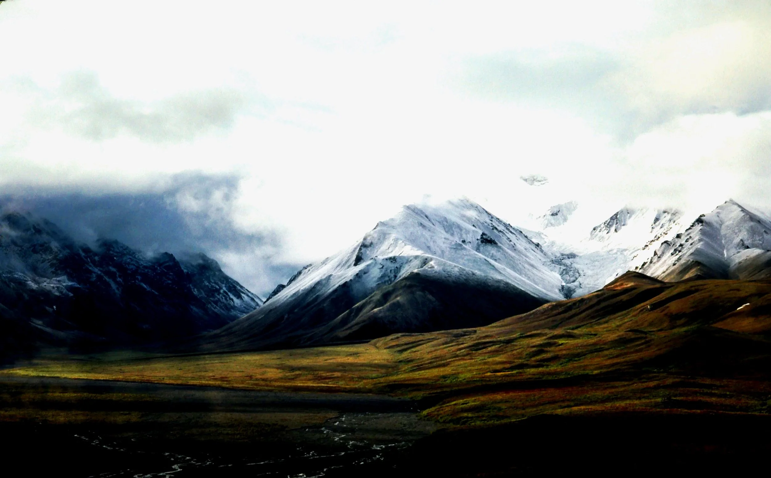 Snow-capped mountains under a cloudy sky with a grassy valley in the foreground.