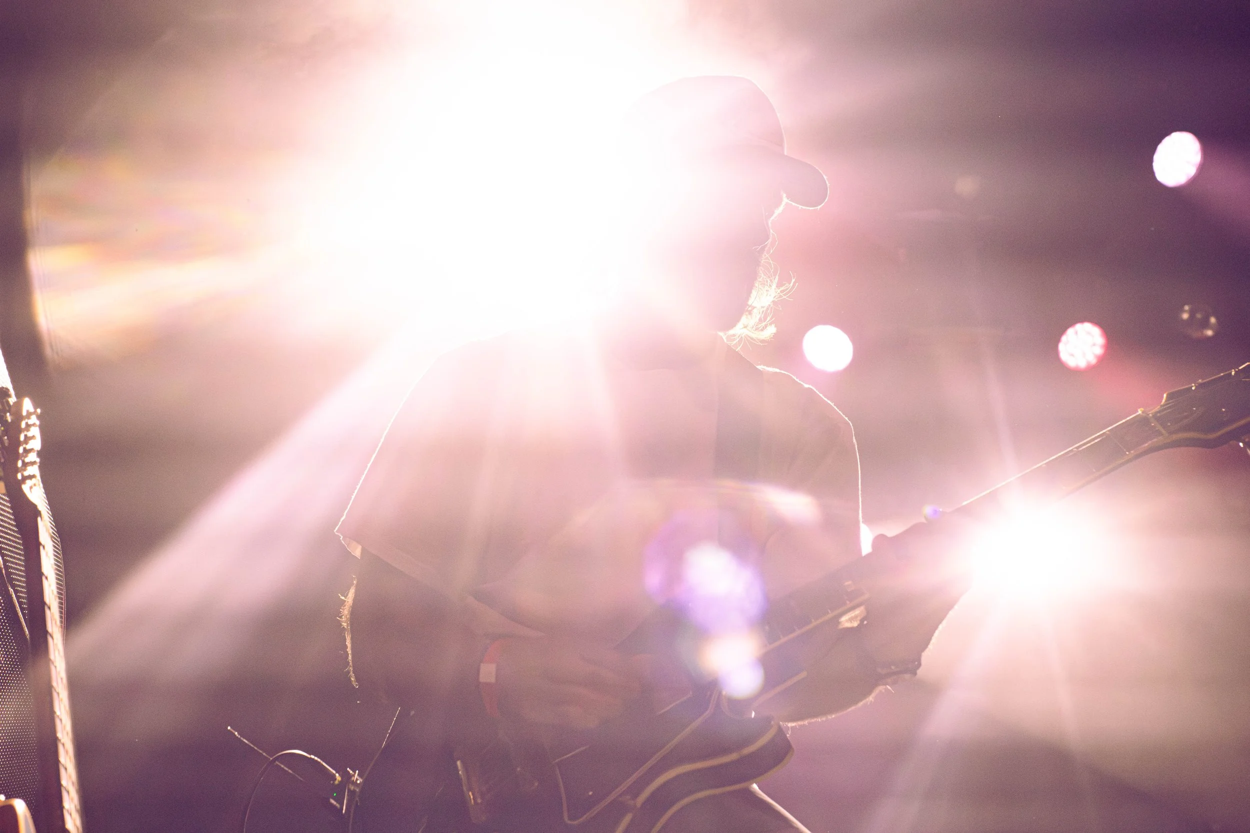 Silhouette of a person wearing a hat, possibly a musician, illuminated by bright stage lights from behind at a concert or performance.
