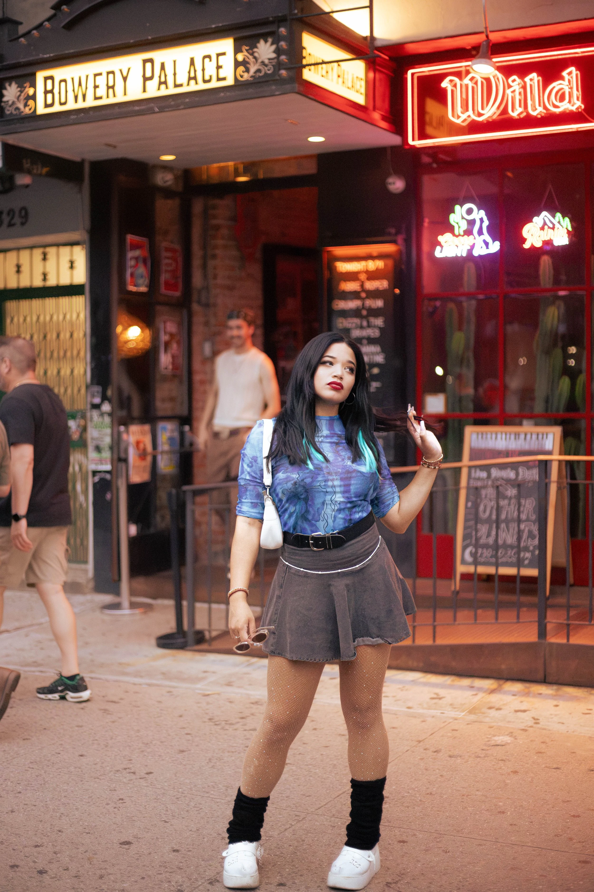 A young woman with long black hair and blue-green streaks stands on a city sidewalk holding sunglasses and smoking a cigarette, with neon signs and brick building in the background. Deanna Valentine Music Performance at Bowery Palace/ Bowery Electric