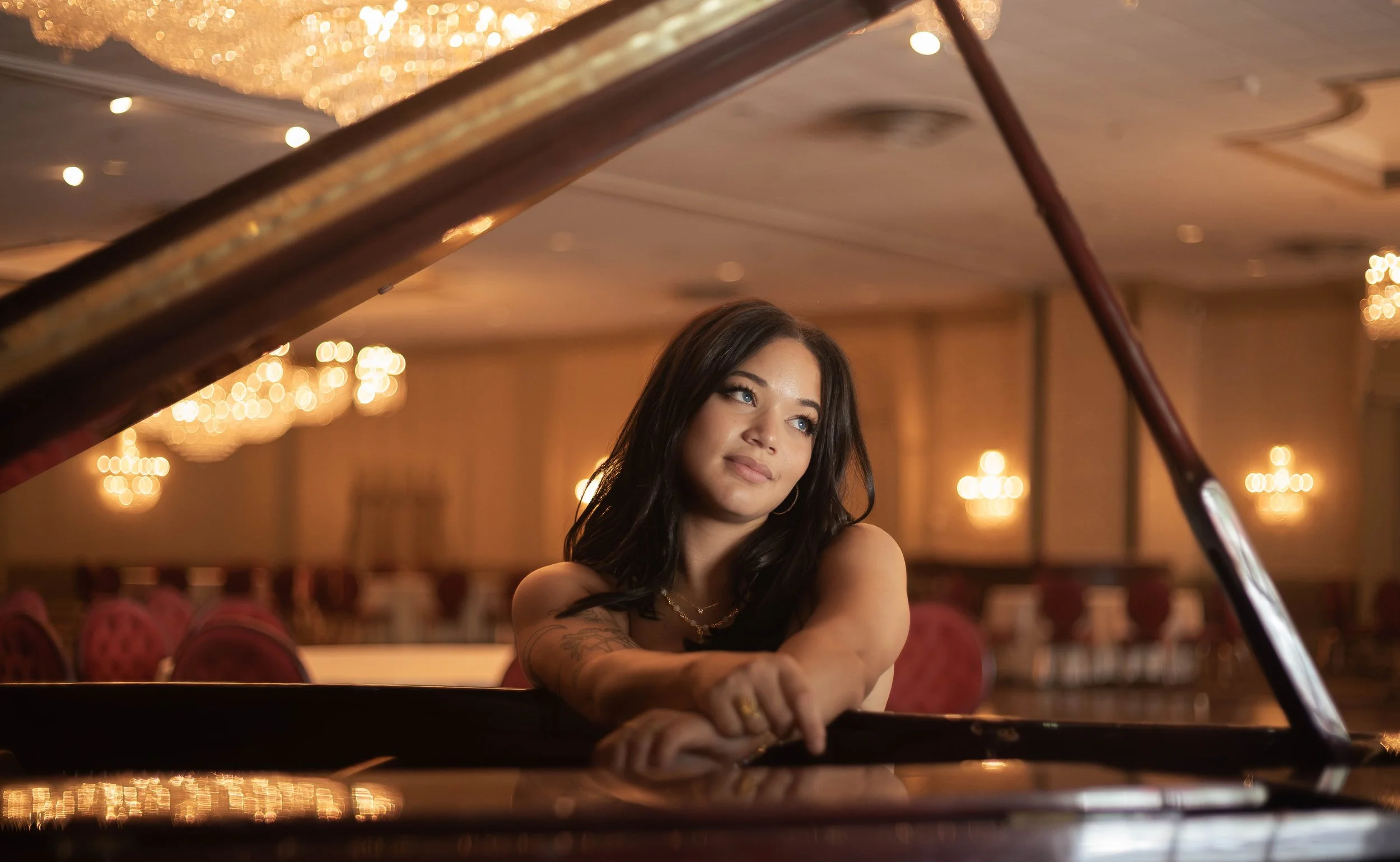 A young woman with dark hair playing the piano in a room decorated with warm lighting and chandeliers.