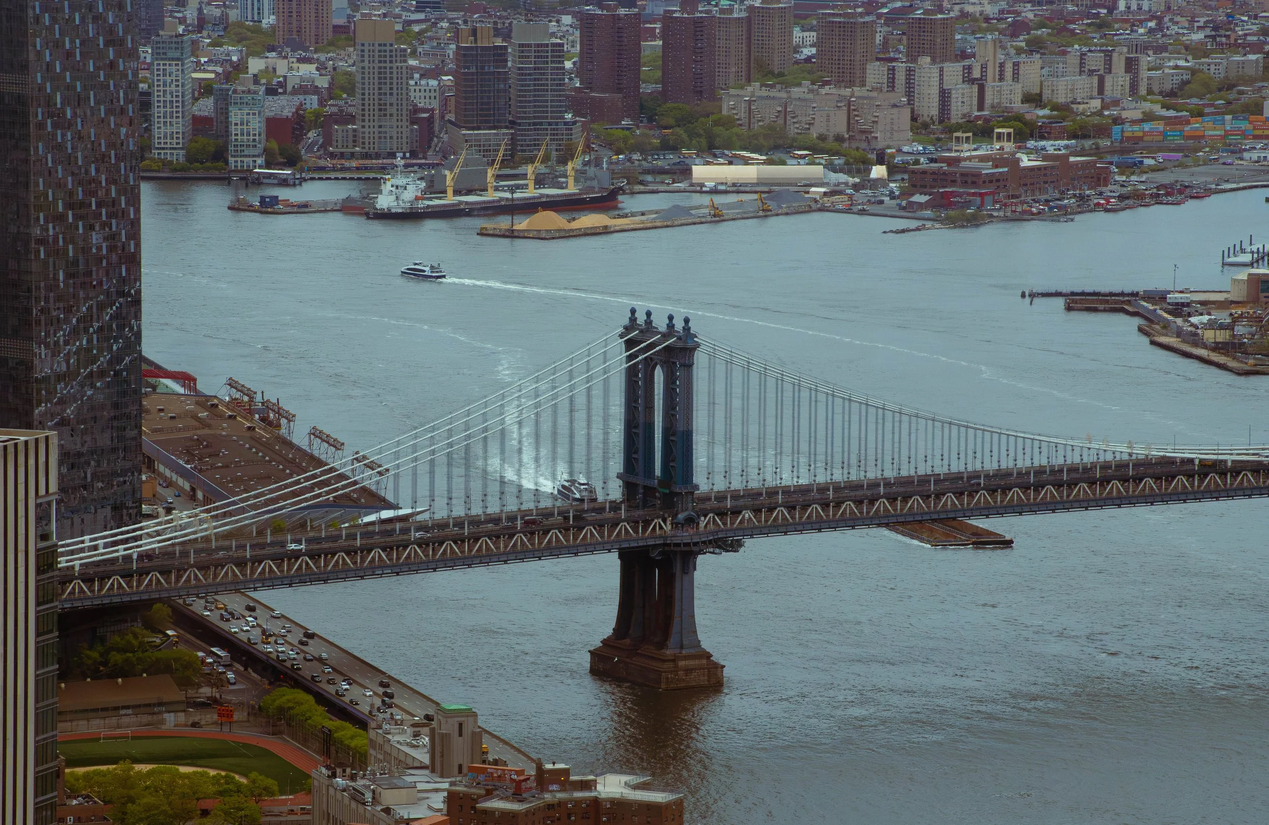 Aerial view of the Manhattan Bridge crossing the East River, with Manhattan skyline and city buildings in the background, and a boat sailing on the water.