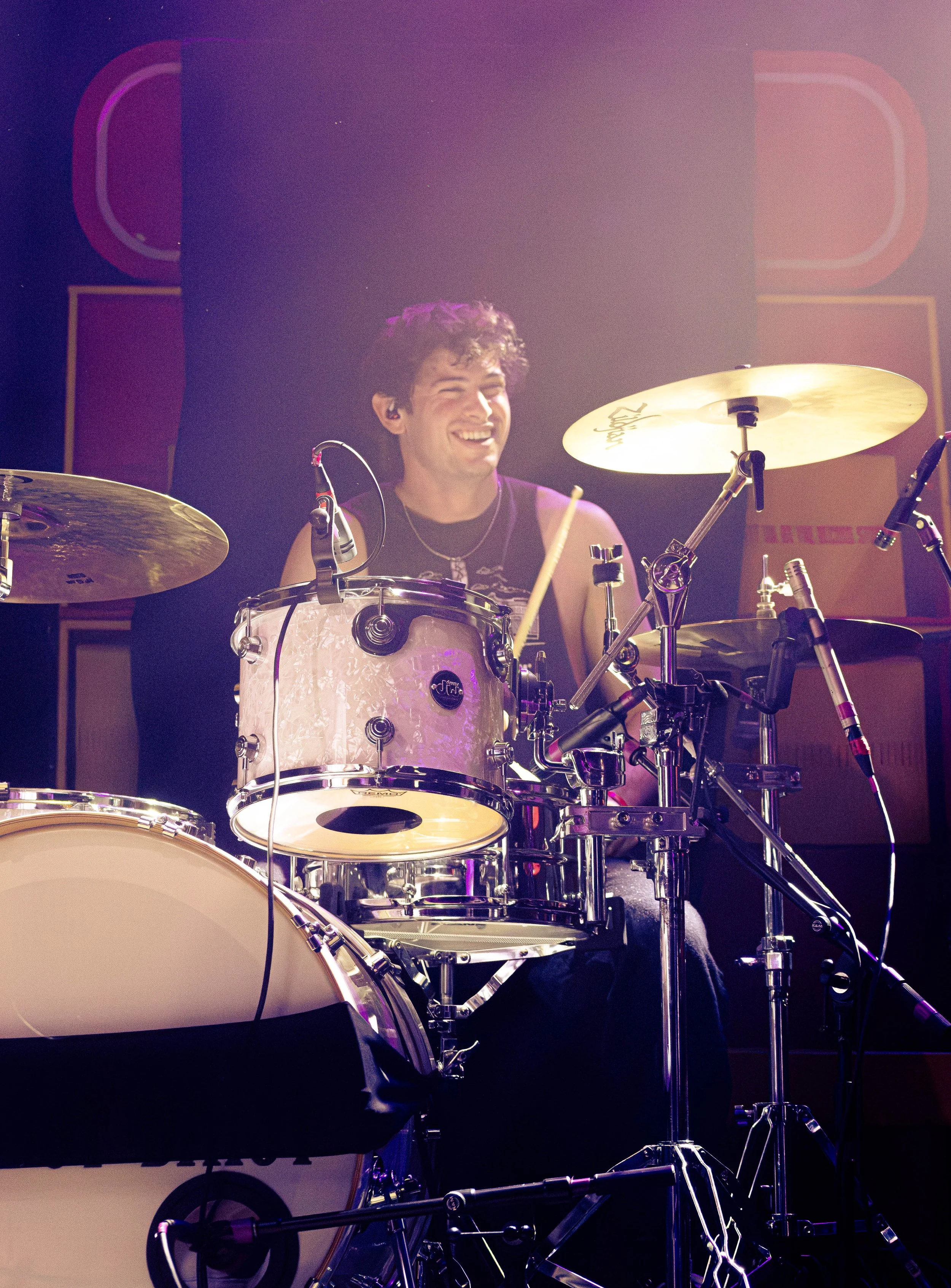 A young man with curly hair and a black sleeveless shirt is smiling and playing a drum set on stage, with a dark background and stage lighting.