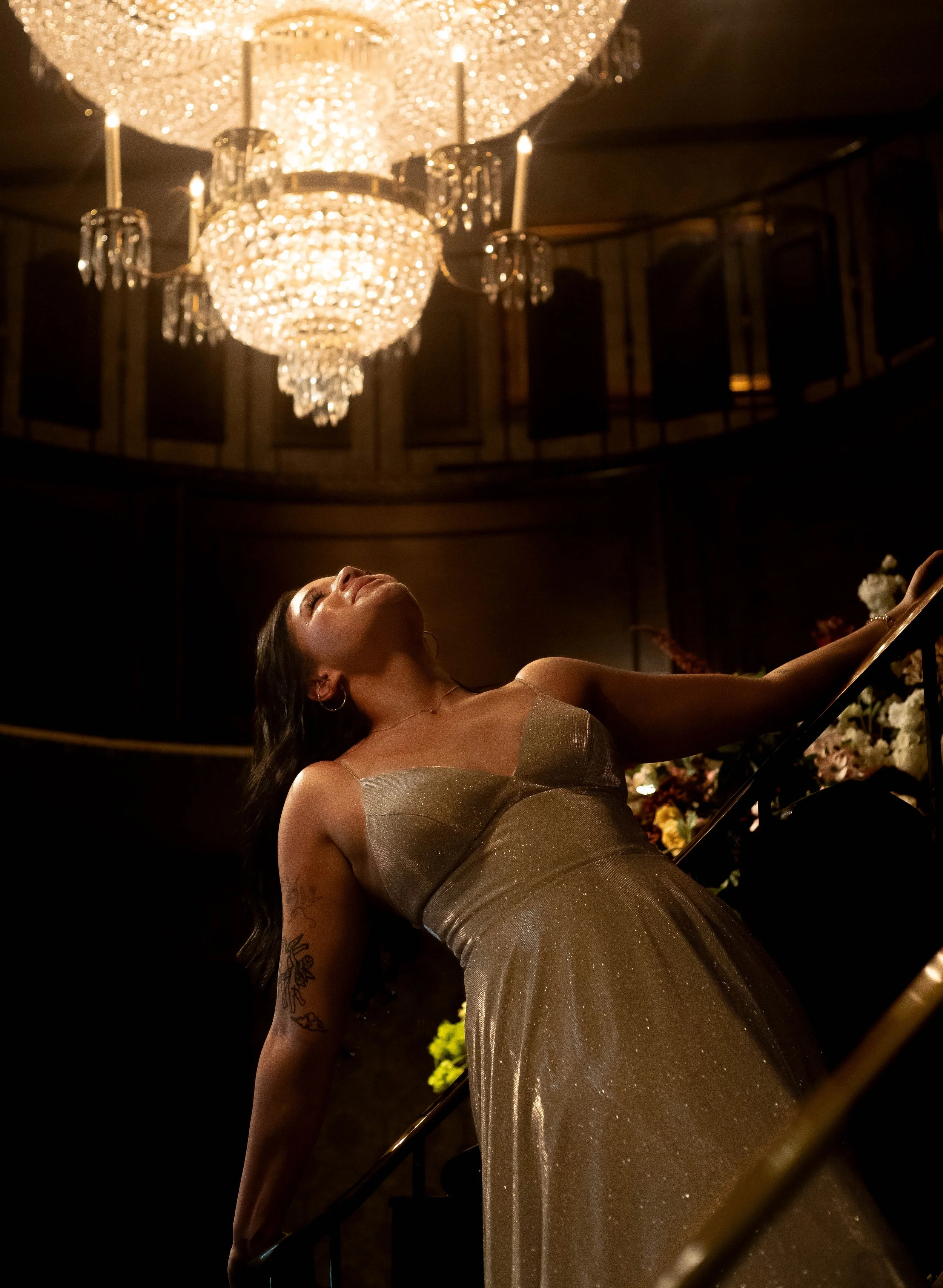 A woman in a shiny gold dress is lying on a staircase, with a chandelier hanging above her in a dimly lit room.