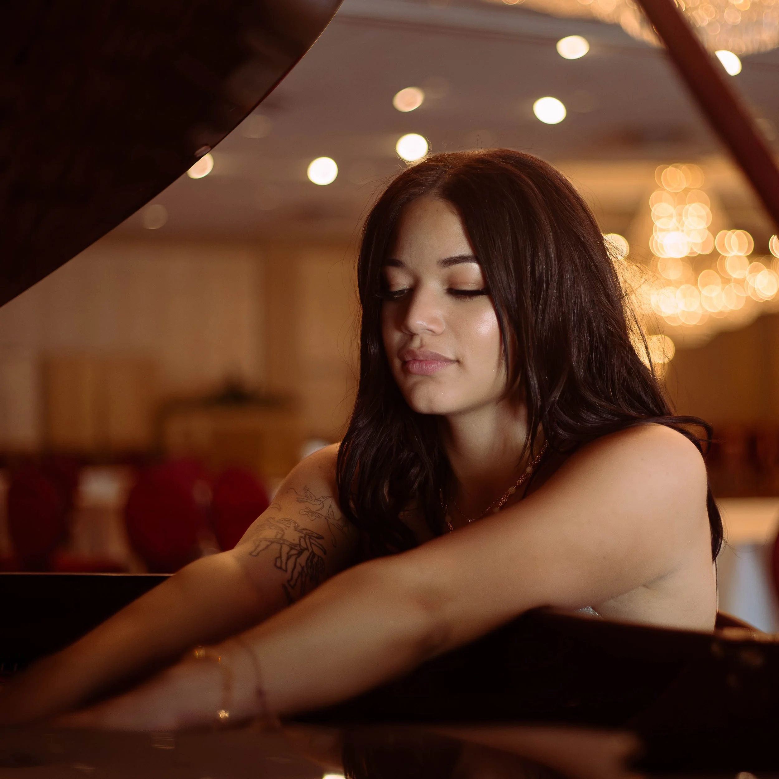 Close-up of a woman with long dark hair and tattoos on her arm, with blurred warm string lights in the background.