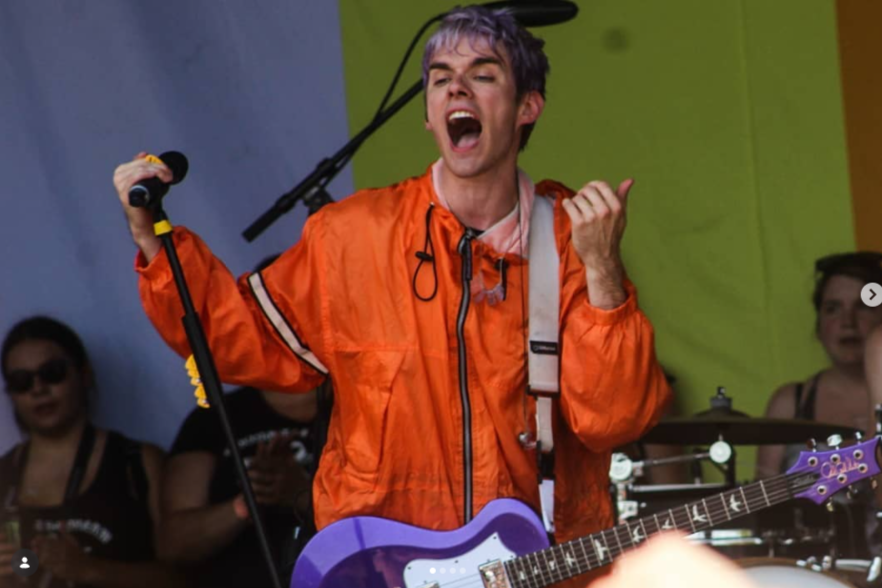 Awsten Knight of Waterparks at Warped Tour 2018 singing passionately into a microphone while holding an electric guitar, wearing an orange jacket, with a colorful background and band members in the background. photo by Annelise Mirabal