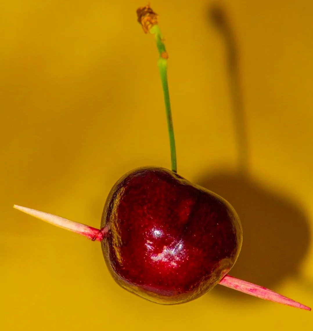 A cherry with a toothpick stuck through it and a green stem, set against a yellow background. Waterparks band inspired album cover by Annelise Mirabal