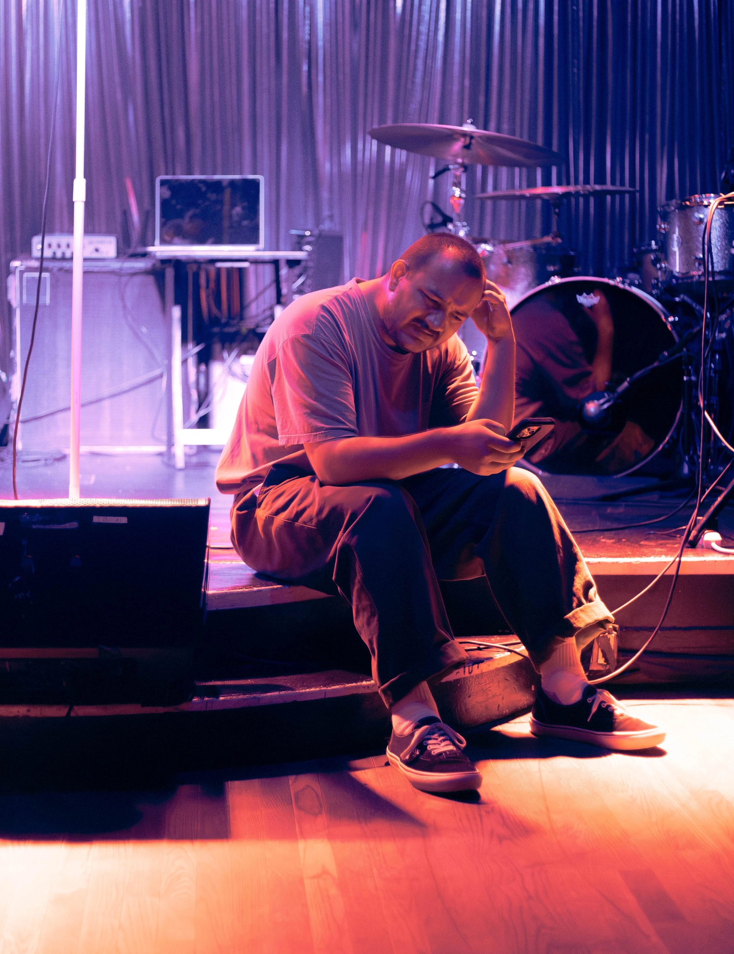 A man sitting on a stage floor in front of musical instruments, looking at his phone with a distressed expression, under purple and orange lighting. Deanna Valentine Music Performance at Bowery Palace/ Bowery Electric on August 11 2026
