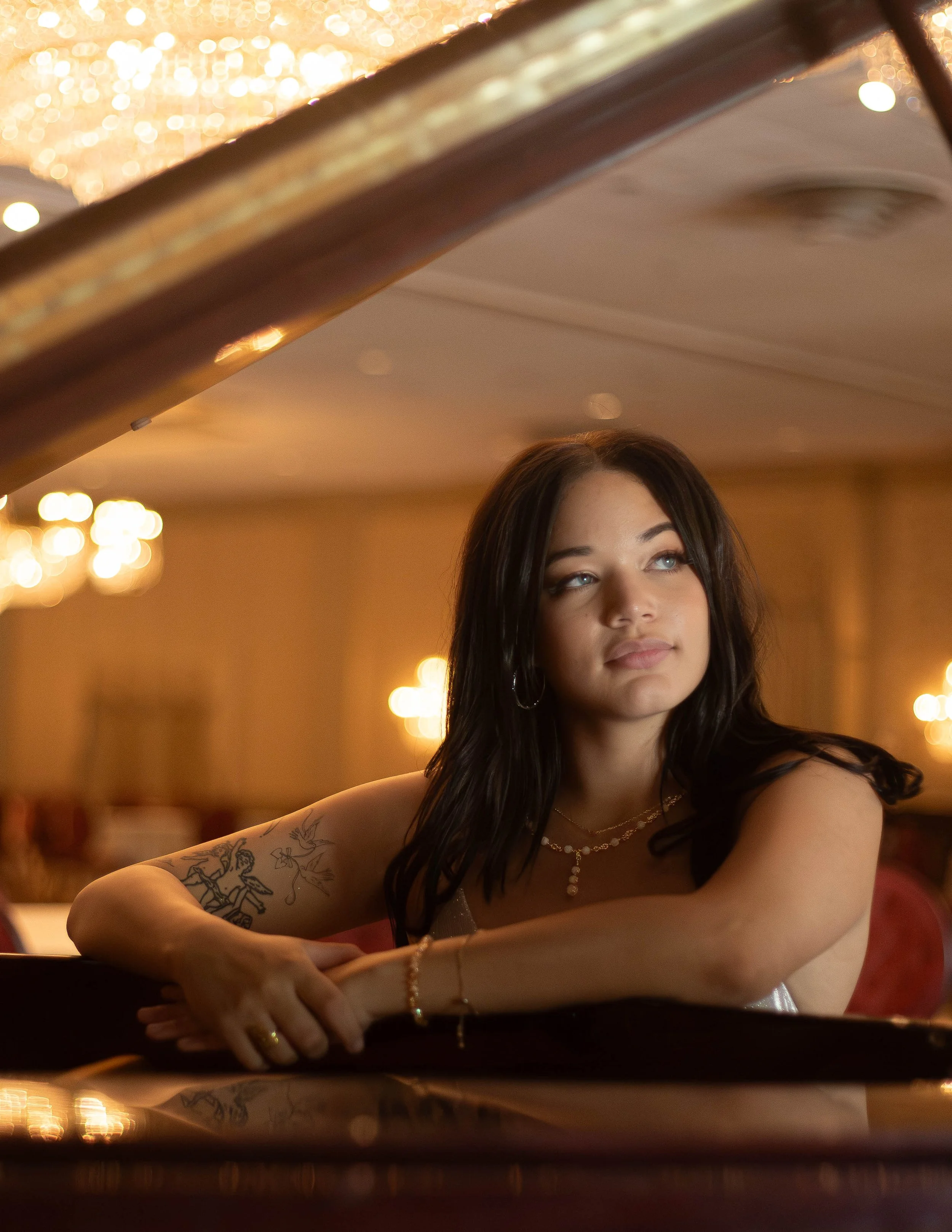 A woman with dark hair and tattoos on her arm sitting at a piano in a warmly lit room with chandeliers.