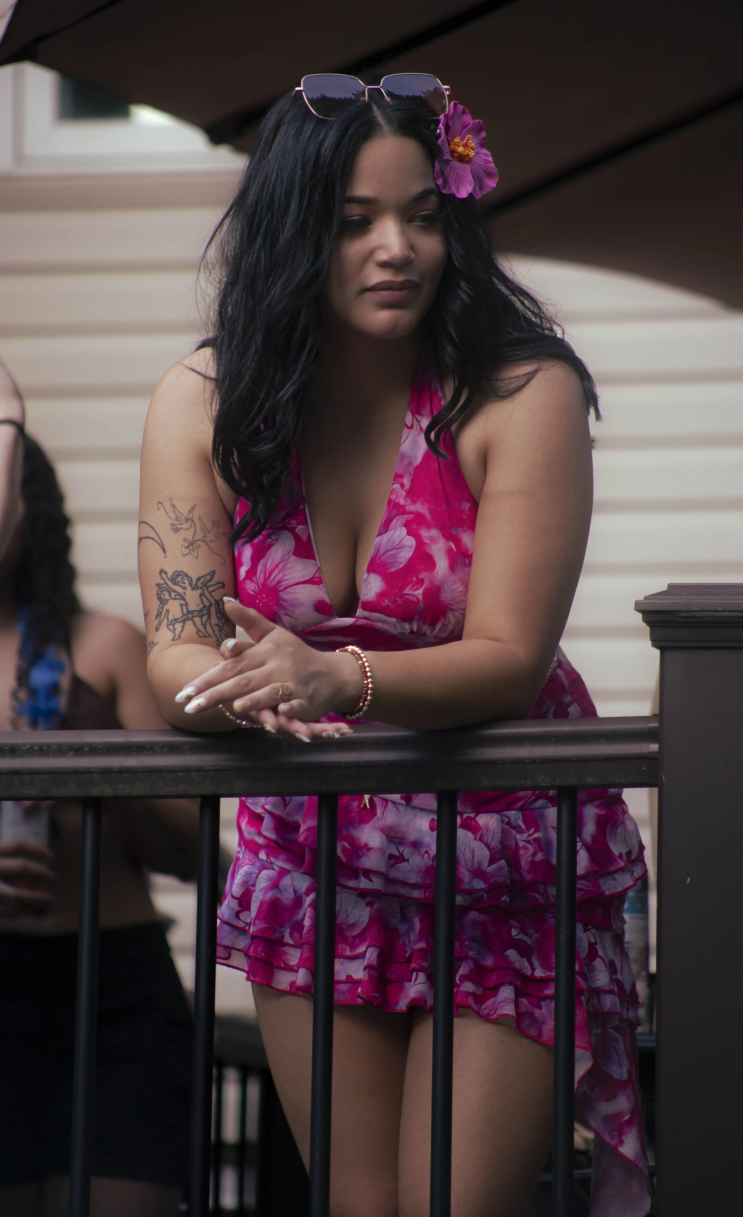 A young woman with long black wavy hair, wearing a pink floral dress, standing behind a black railing on a balcony. She has a purple hibiscus flower in her hair, sunglasses on her head, and tattoos on her left arm. She is looking to her left with a neutral expression.