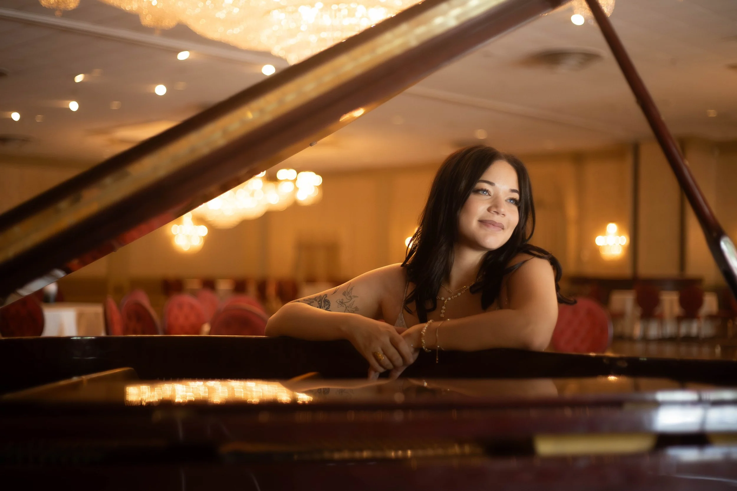 A young woman with dark brown hair and tattoos on her arm, sitting at a grand piano in an elegant, warmly lit room.