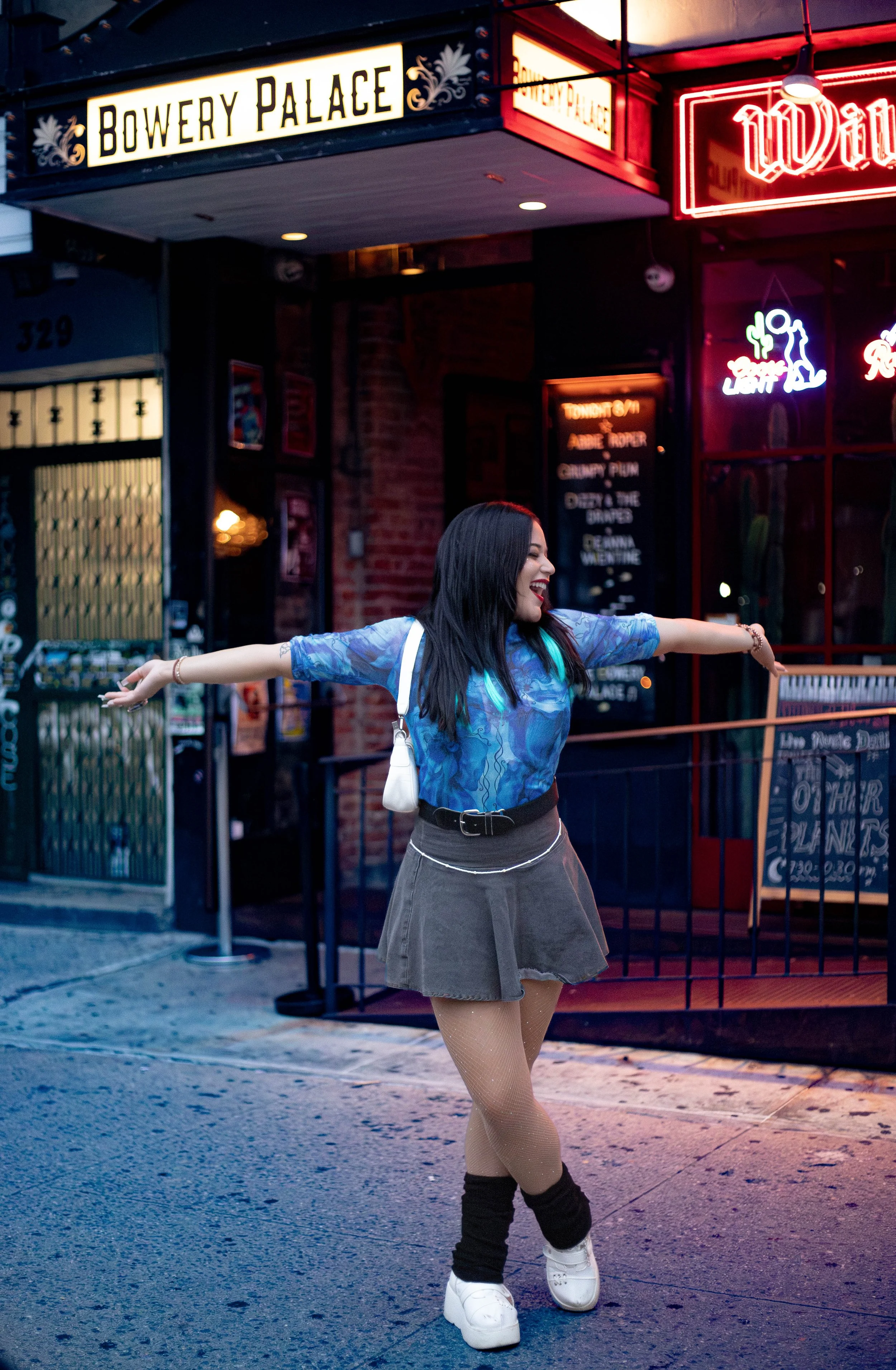 A woman with dark hair, smiling and wearing a blue top, a brown skirt, fishnet stockings, and white sneakers, dancing on the sidewalk in front of a restaurant called Bowery Palace with neon signs, including one for Budweiser and a neon sign with a gi