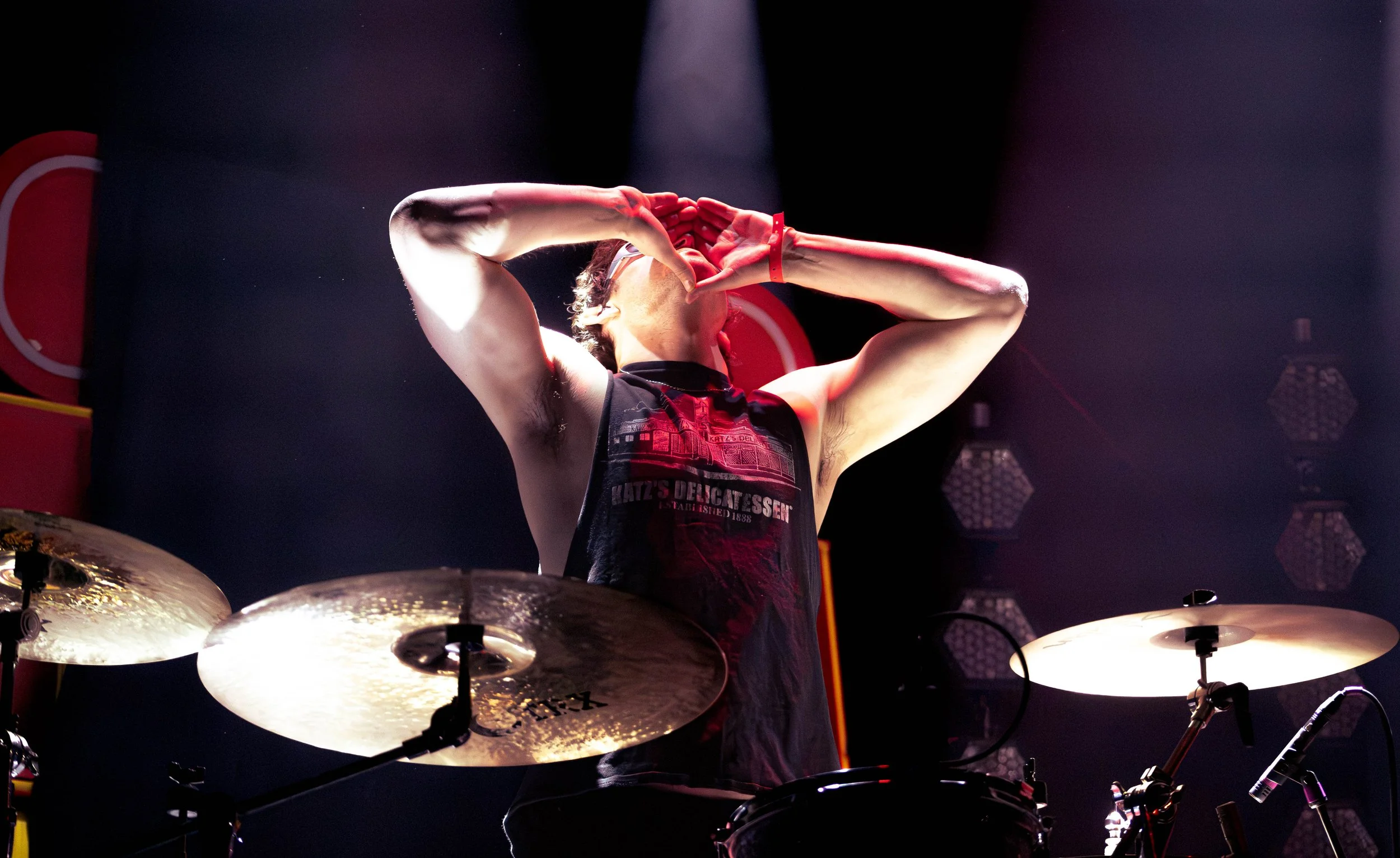 A drummer playing drums on stage, seen from below with cymbals in the foreground, stage lights illuminating the scene, and a dark background.