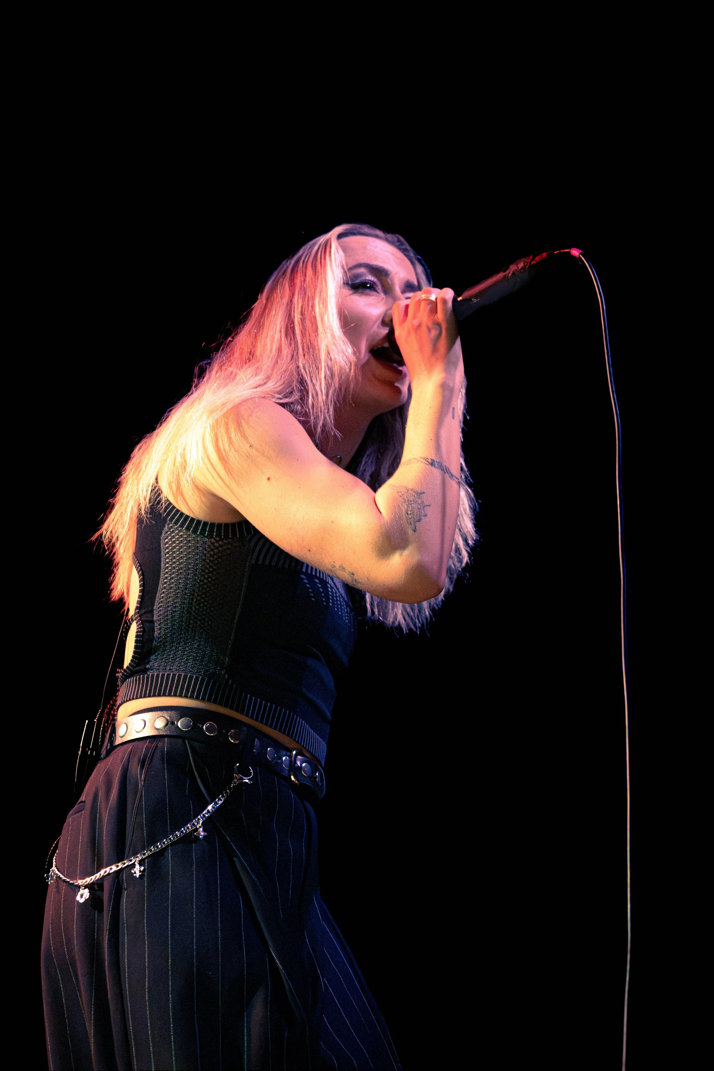 A female singer with long blonde hair performing on stage, holding a microphone and singing passionately. She is wearing a black sleeveless top and black pinstripe pants with silver chains.