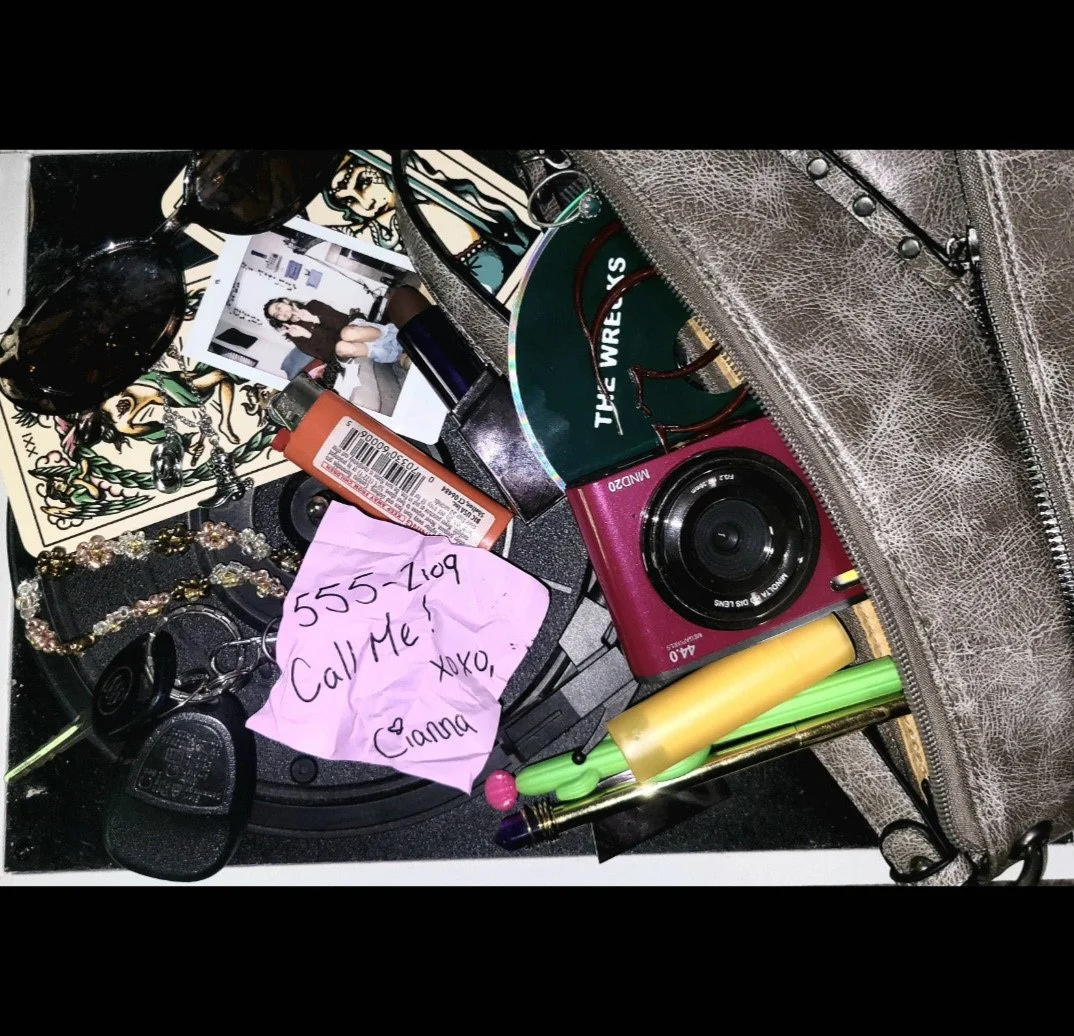 Cluttered jewelry and cosmetics bag containing sunglasses, a Polaroid photo, a pink camera, a yellow and green highlighter, a pink sticky note with phone number and message, lip gloss, a bracelet, and other small items.
