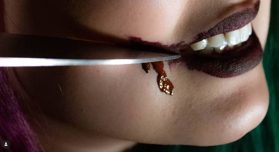 Close-up of a person with makeup and lipstick, smiling slightly, with a knife holding a viscous brown substance near their chin. photo by Annelise Mirabal