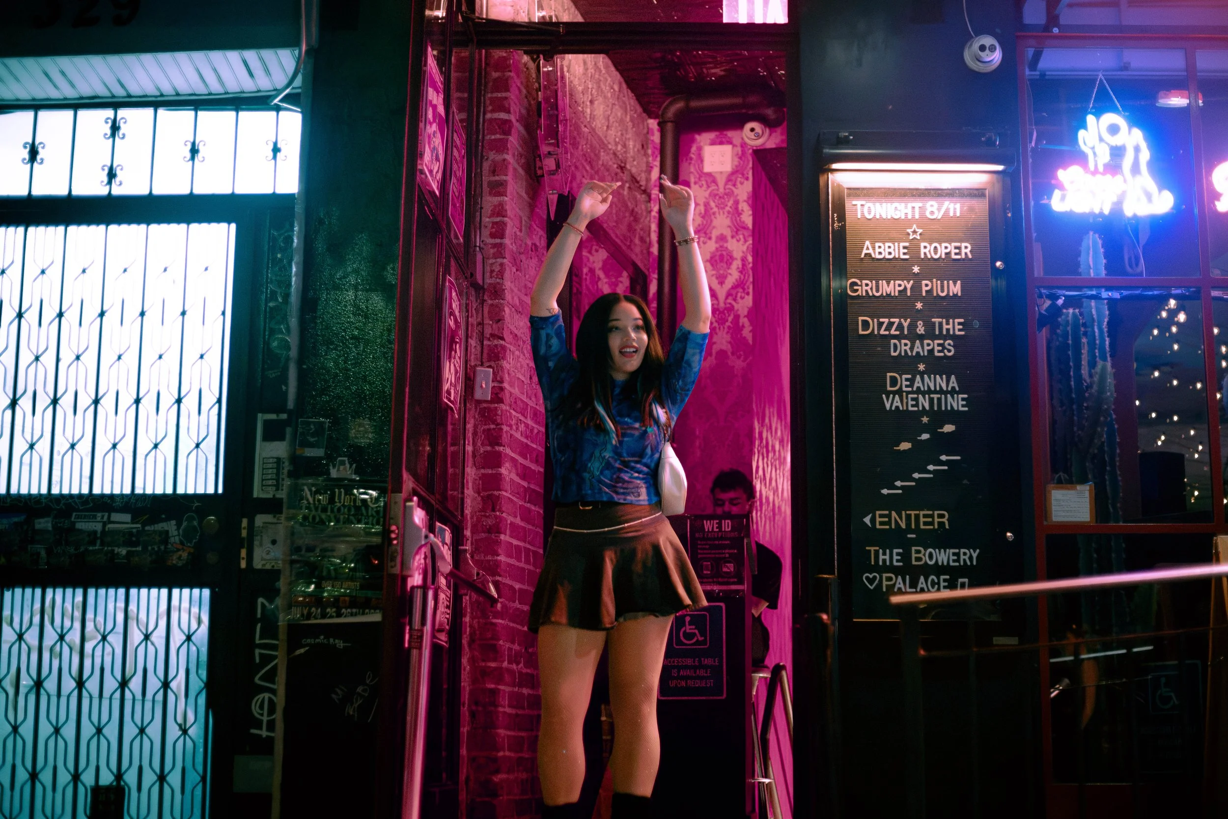 A young woman with long dark hair, wearing a blue patterned top and a black skirt, standing at the entrance of a bar or club with pink and purple neon lighting, raising her hands in the air and smiling. Deanna Valentine Music Performance at Bowery Pa