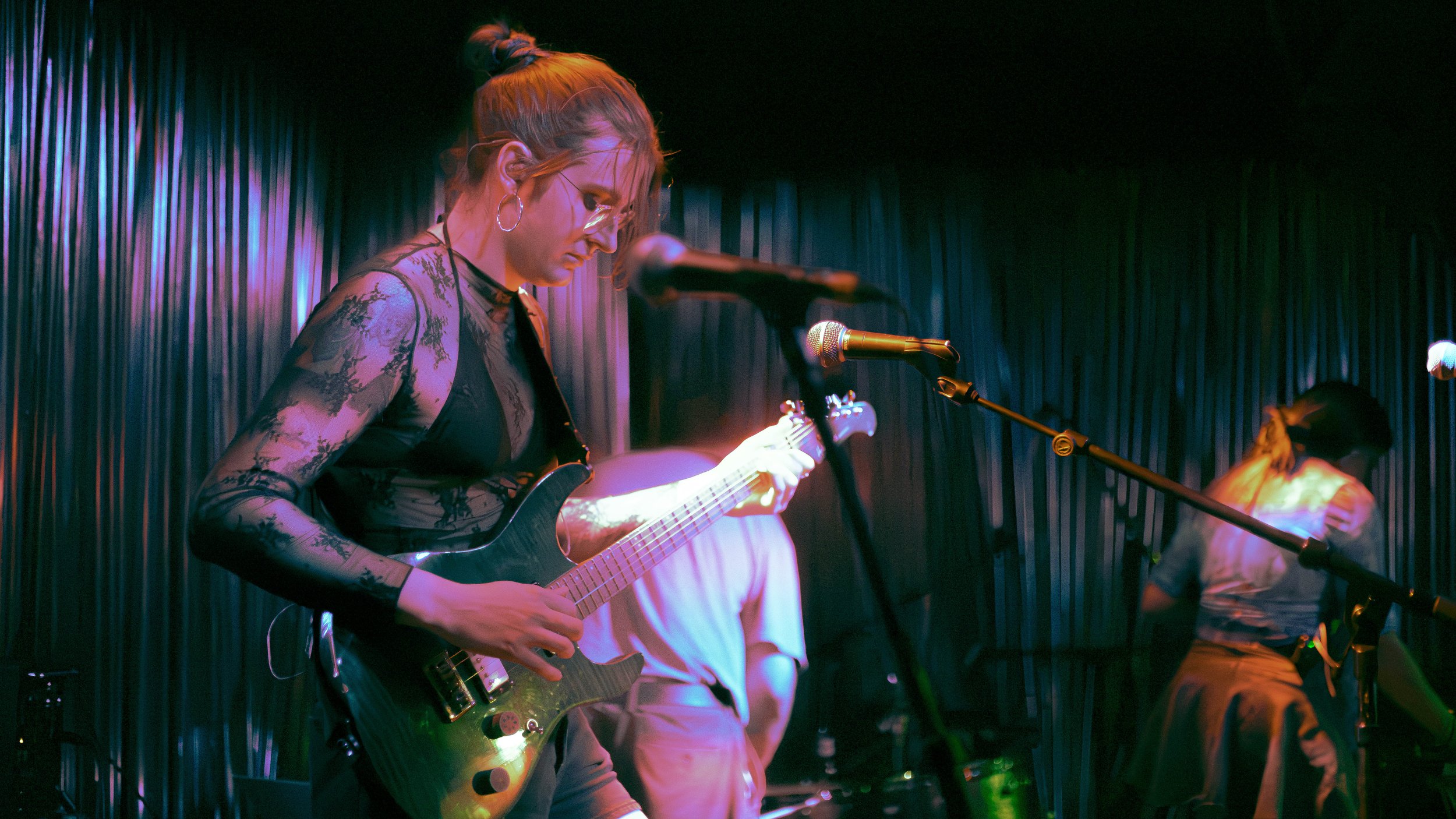 A woman playing an electric guitar on stage with a dark curtain background, wearing glasses and a black lace top, illuminated by colorful stage lighting. Deanna Valentine Music Performance at Bowery Palace/ Bowery Electric on August 11 2026