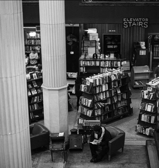 A man reading a book in a bookstore sitting on an armchair. There are bookshelves filled with books surrounding him, and a large column is in the foreground. Signage indicating elevator and stairs is visible in the background.