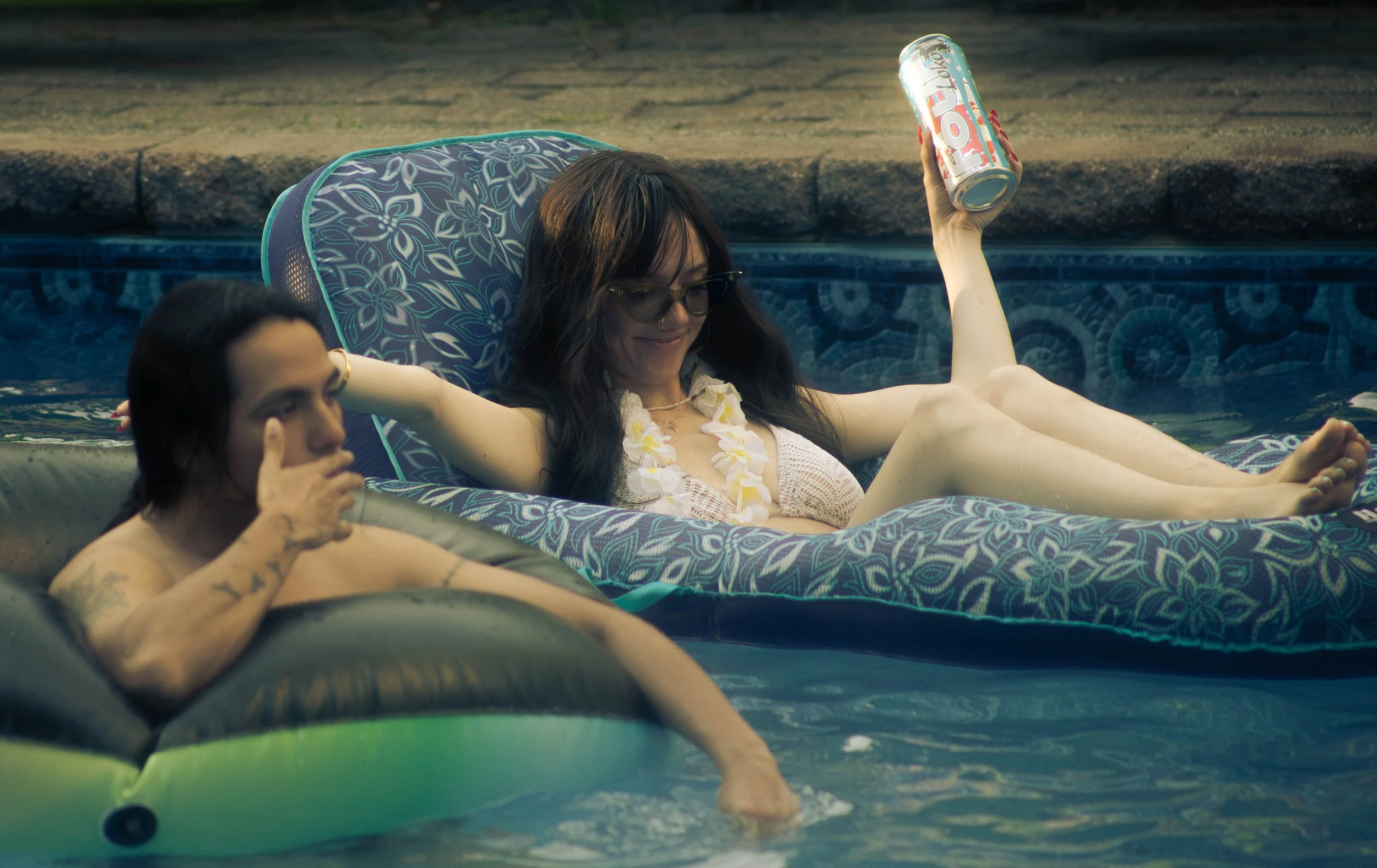A woman with glasses and dark hair relaxing on an inflatable pool float, holding a can of Fanta soda, with a person next to her in a pool.