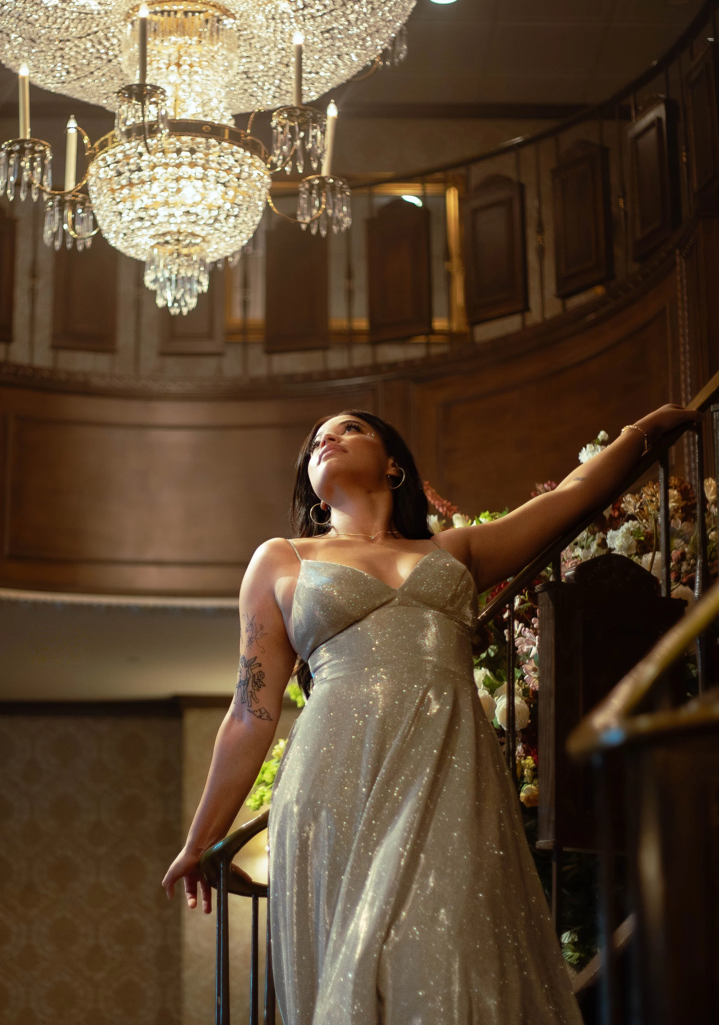 A woman in a shimmering, light-colored dress standing on a staircase, with her arm extended and looking upward, under a large, ornate crystal chandelier in a grand, wood-paneled interior.