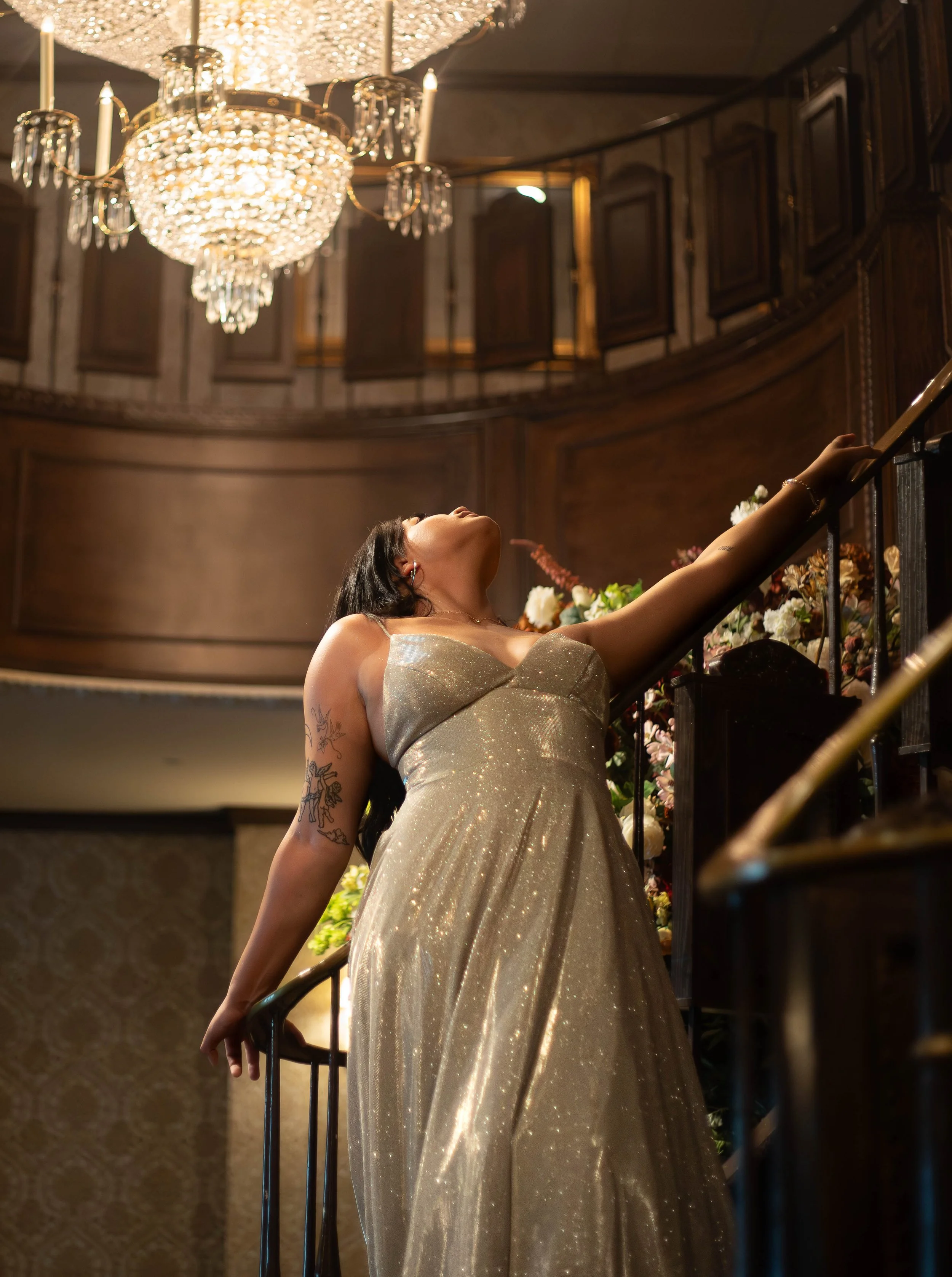 A woman in a shimmering gold gown is ascending a staircase, reaching for the handrail, with a large crystal chandelier hanging above and a wooden-paneled balcony in the background.