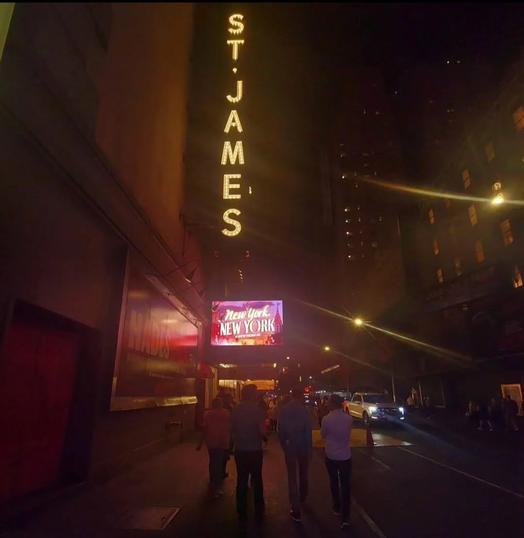Night street scene with illuminated 'ST. JAMES' sign, digital billboard displaying 'New York,' and people walking on the sidewalk.