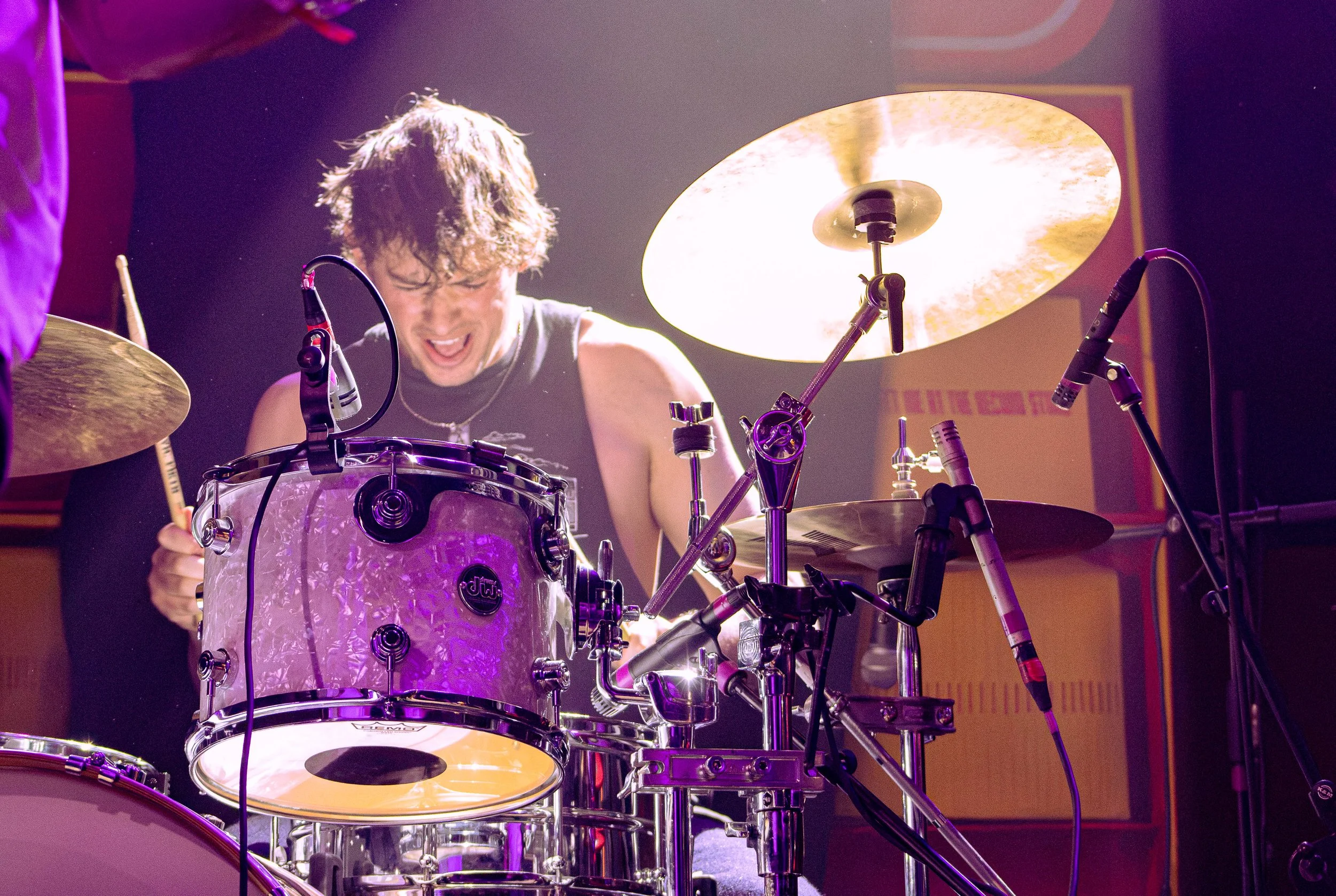 A musician playing a drum set on stage, with a focused expression, illuminated by colorful stage lights.