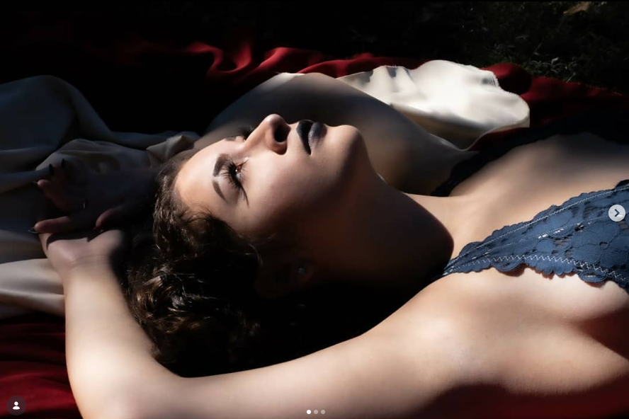 A woman lying on her back with her eyes closed, wearing a lace top, surrounded by soft fabrics in dark and light colors.