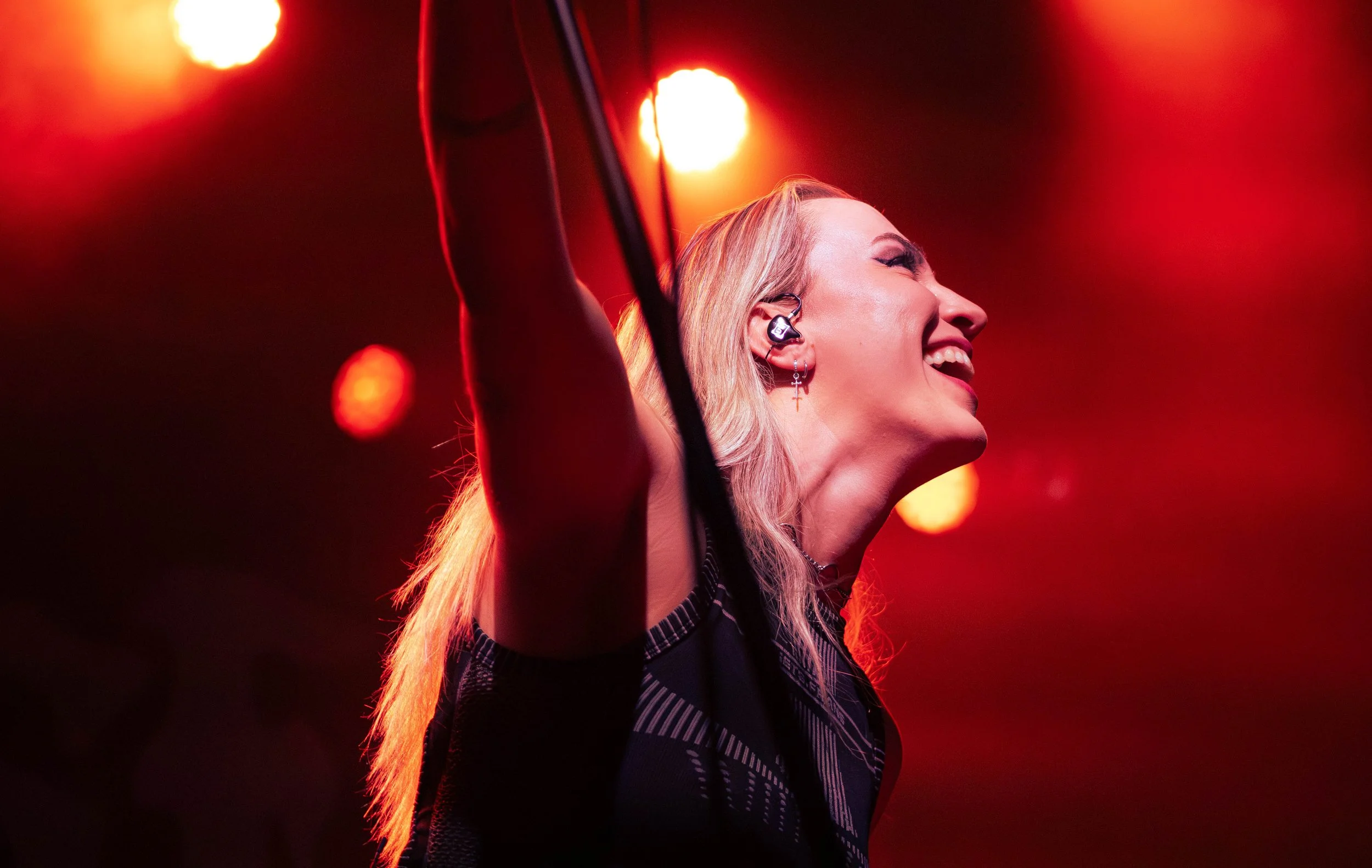 A woman with long blonde hair smiling on stage, illuminated by red and orange stage lights, wearing an earpiece and dark clothing.