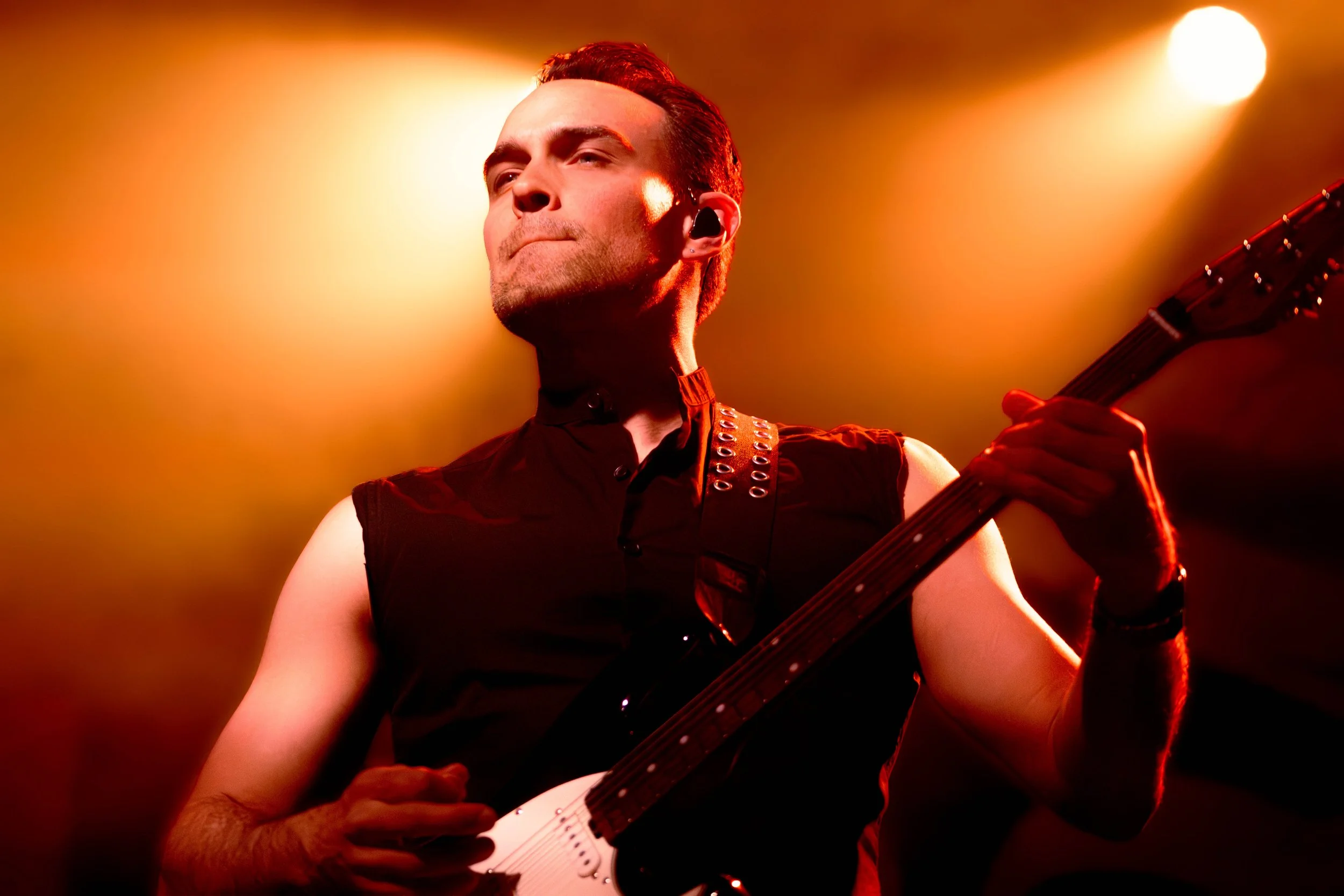 A male performer playing an electric guitar on stage under orange and yellow spotlights.