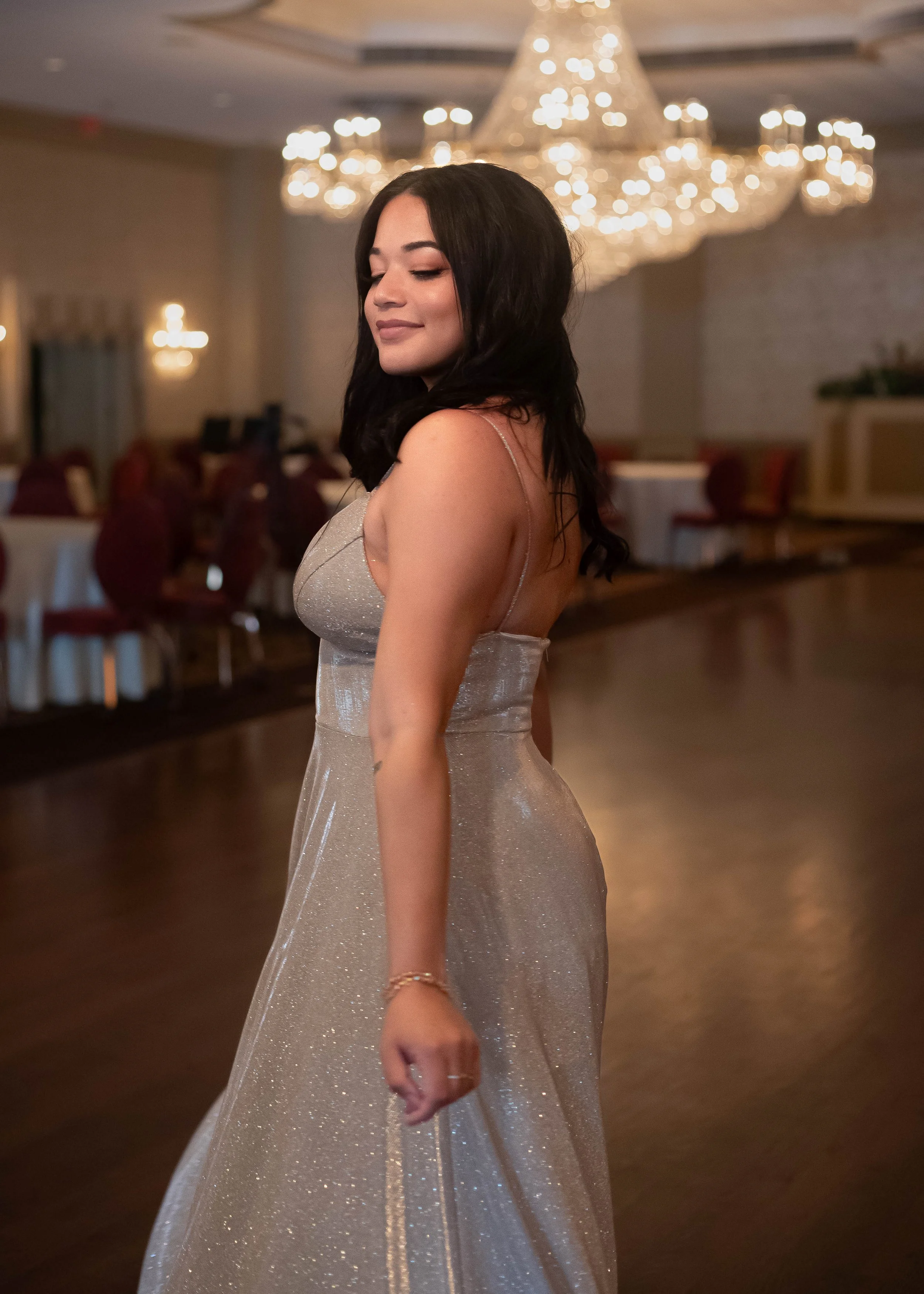 Woman with dark hair wearing a silver, sparkly dress at an indoor event with a chandelier in the background.