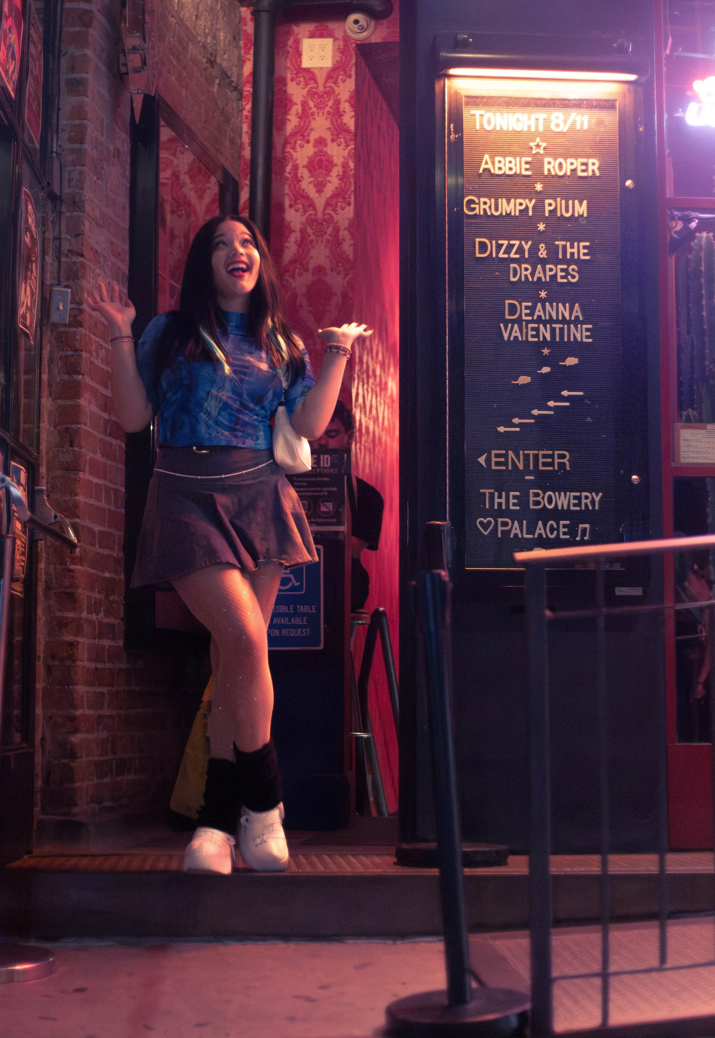 A woman in a blue tie-dye shirt, a brown skirt, and white sneakers stands outside a venue, smiling and waving. A signboard in front lists performers for an evening event, including Abbie Roper and Dizzy & The Drapes. Deanna Valentine Music Performanc