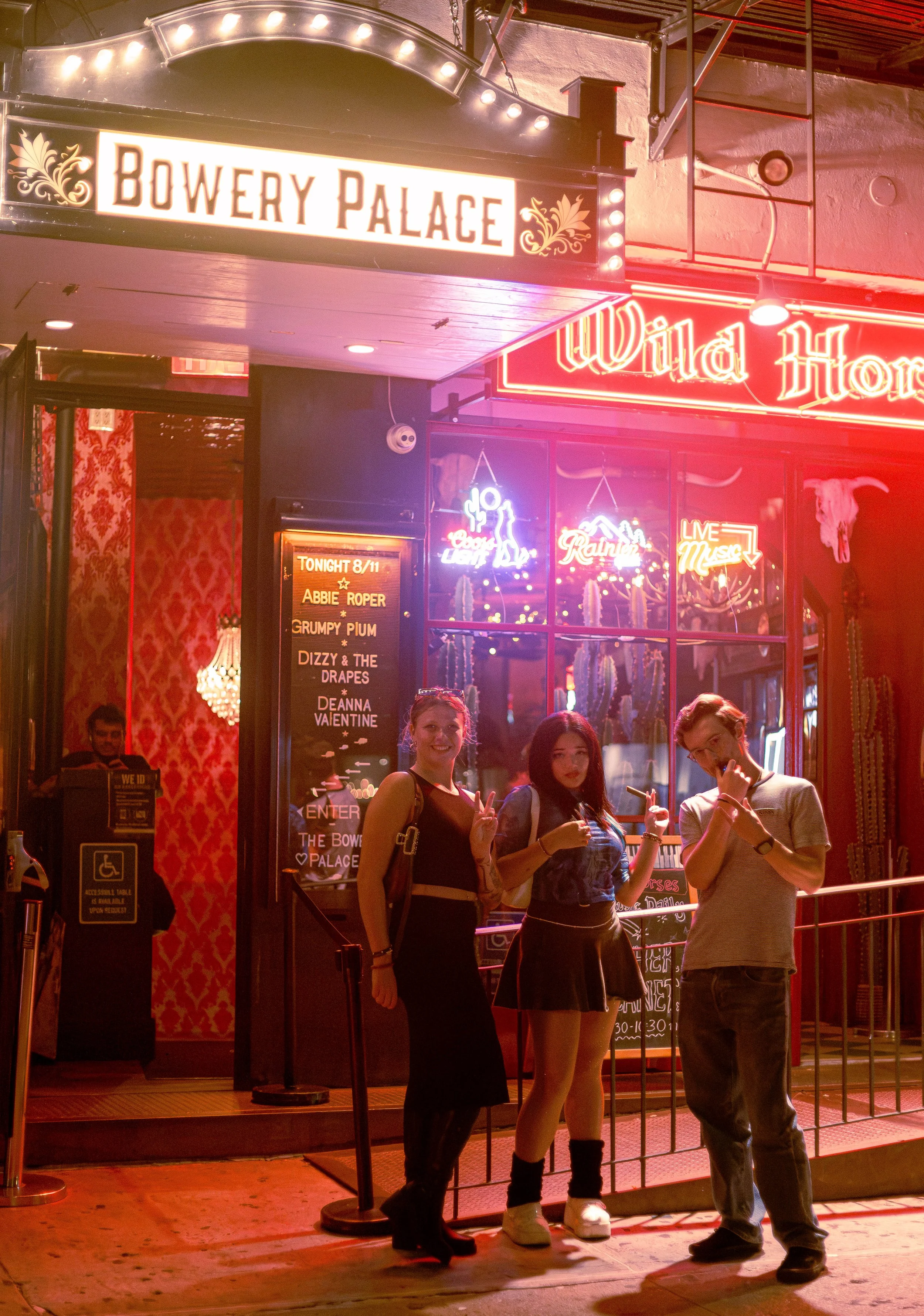 Three young adults standing outside a nightlife venue called Bowery Palace, smiling at the camera with a neon sign for Wild Horse behind them. Deanna Valentine Music Performance at Bowery Palace/ Bowery Electric on August 11 2026