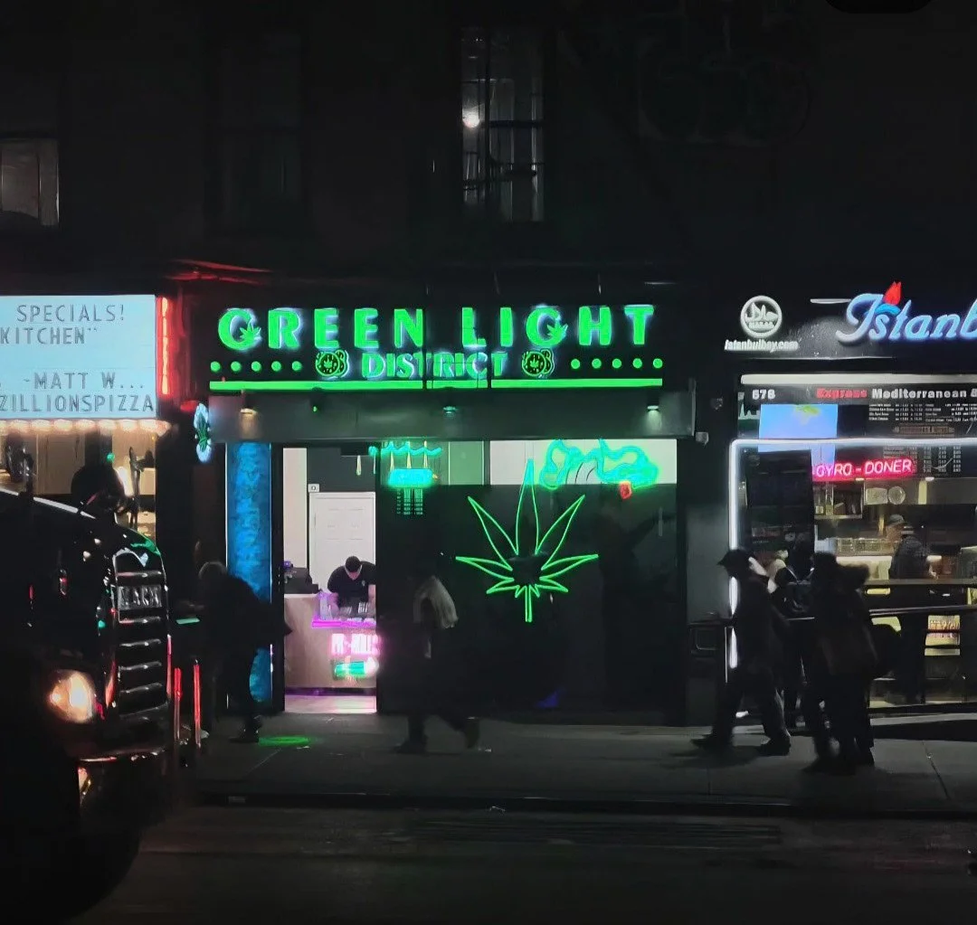 Nighttime street scene featuring a storefront with neon signs, including a large green sign reading 'Green Light District' and a neon marijuana leaf symbol beneath it. Several people are walking on the sidewalk in front, and other storefronts with si