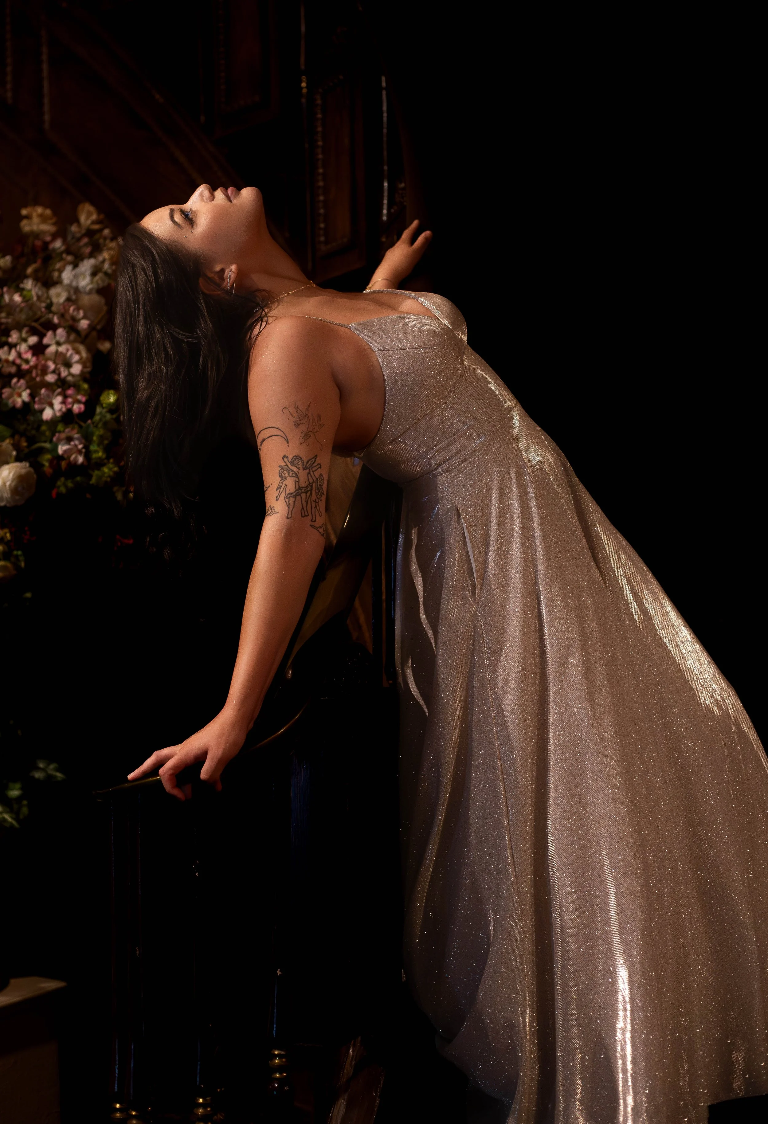 A woman with dark hair and tattoos is leaning back with her eyes closed, wearing a sparkly silver gown, in front of a dark background with pink and white flowers.