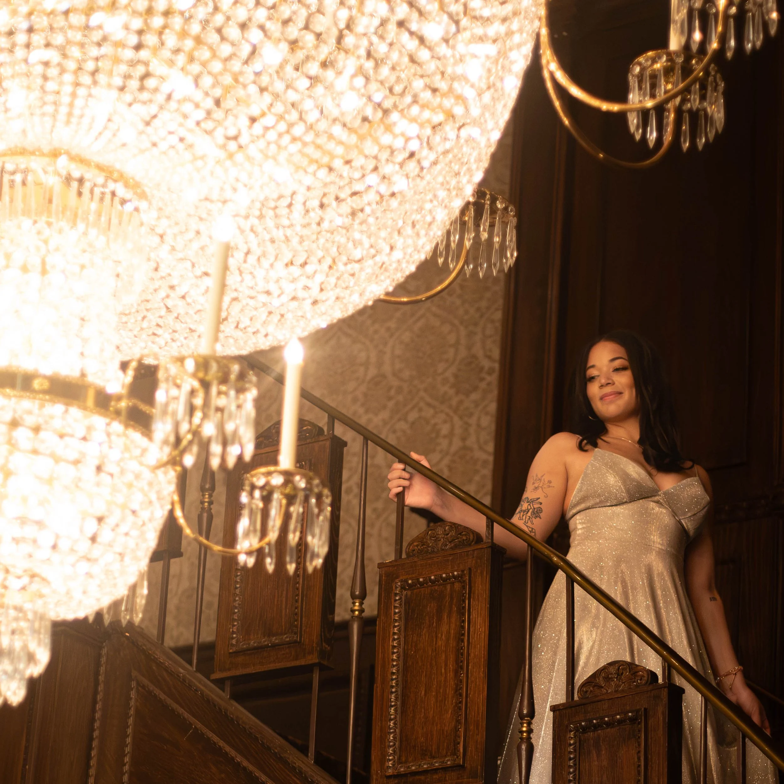 A woman in a gold dress standing on a staircase with ornate woodwork, holding a railing. A large, sparkling chandelier hangs overhead, illuminating the scene.