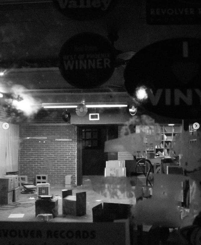 An indoor space with a brick wall and shelves filled with books, boxes, and equipment. The room appears to be a recording or rehearsal studio with dim lighting.