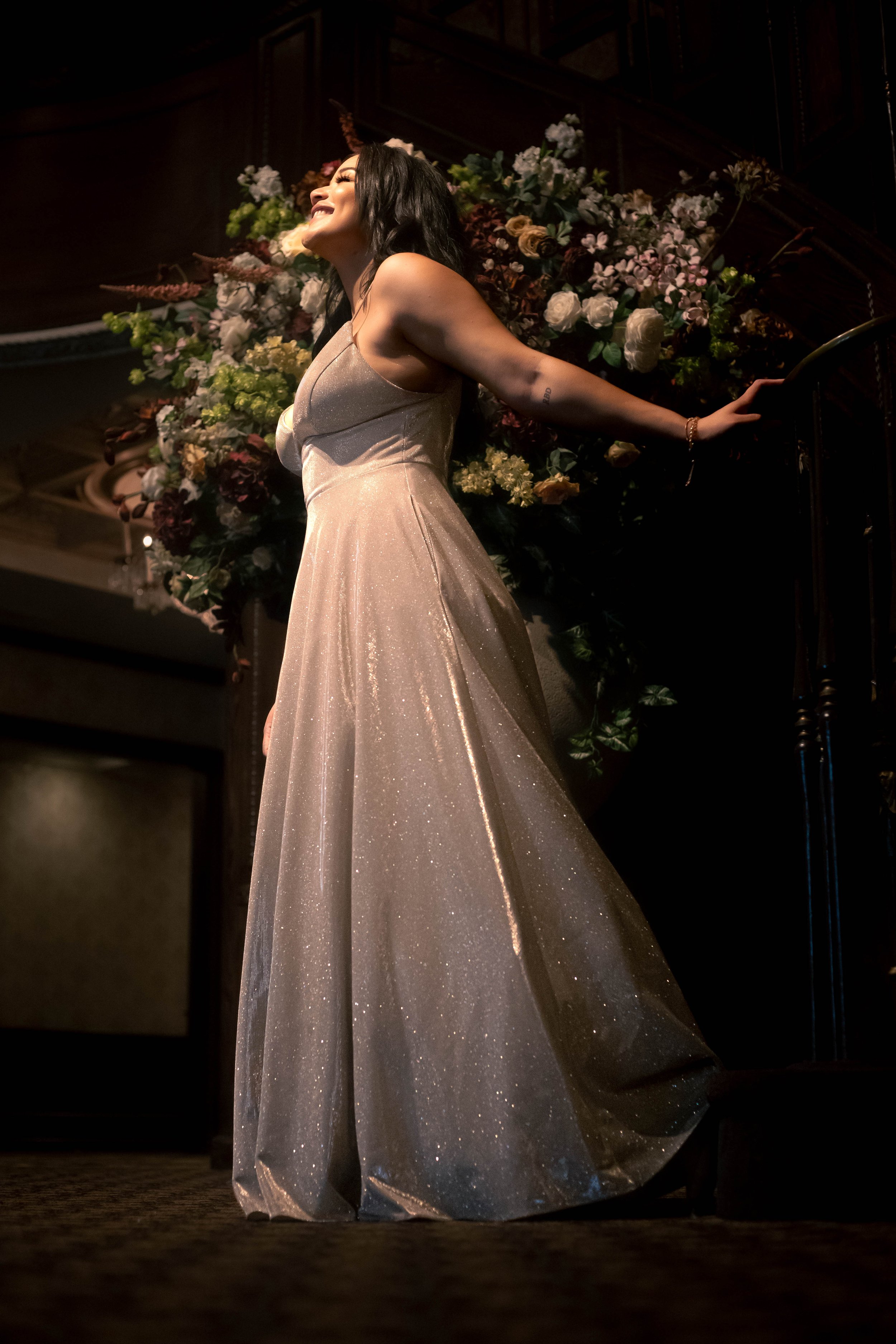 A woman in a sparkling light-colored gown standing on a stage with floral decorations behind her, smiling joyfully, with her arms extended.