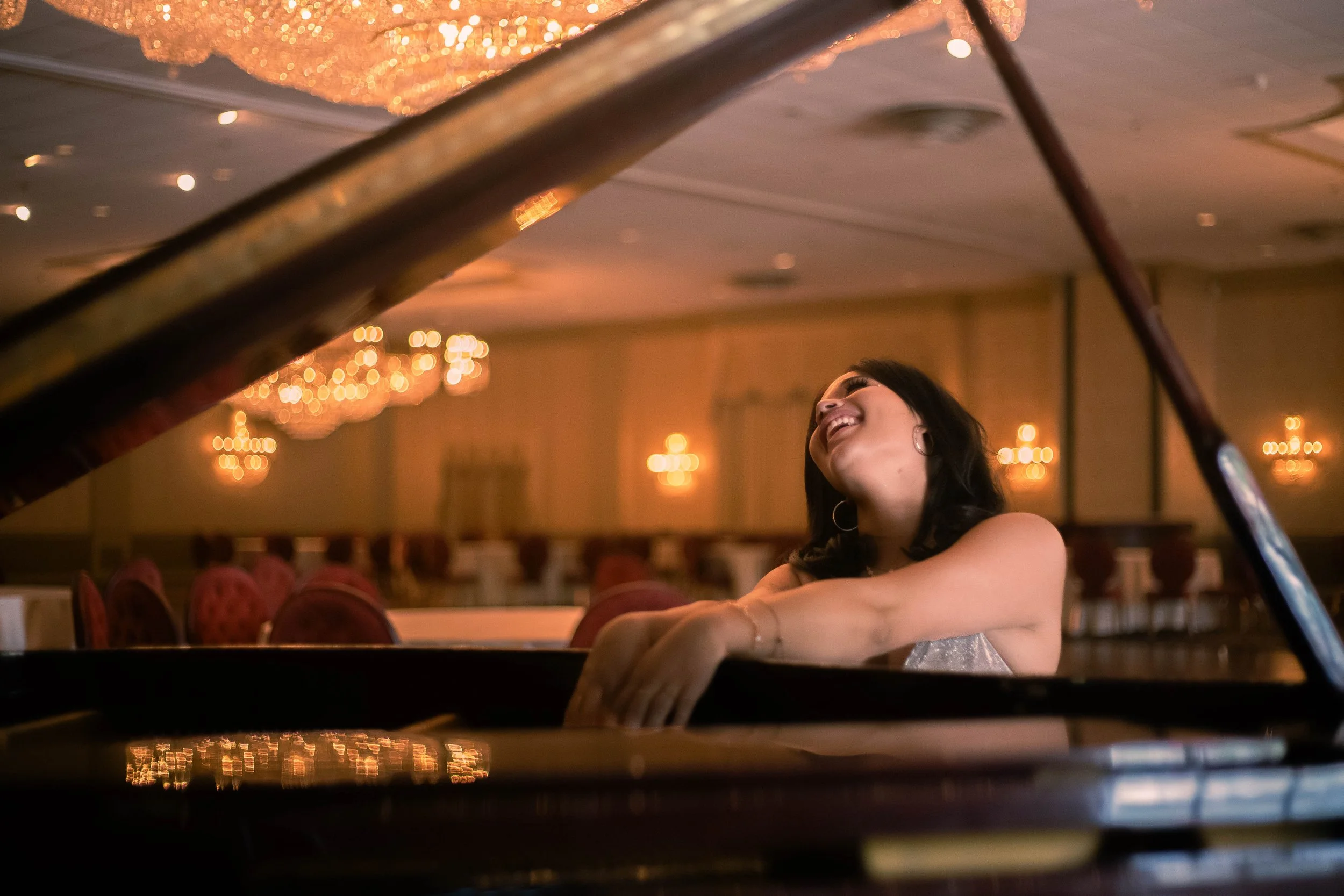 A woman smiling while playing the piano in a warmly lit room with chandeliers and empty chairs.