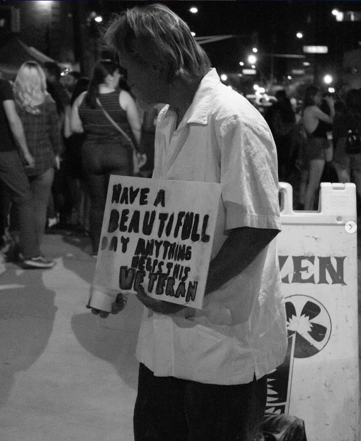 A person holding a sign that reads, "Have a beautiful day. Anything helps. Thank you." standing in a crowd at night.