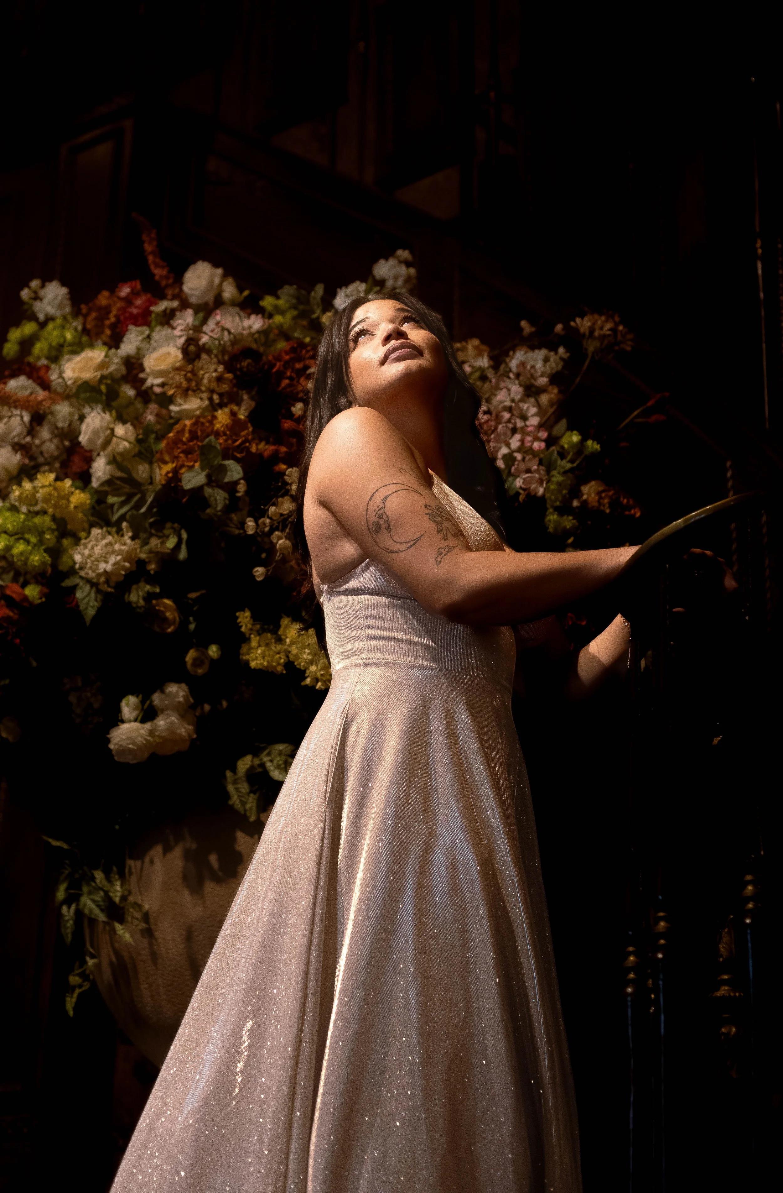 A woman in a pink gown with a glittery texture, holding a railing, standing in front of a backdrop of colorful flowers, looking up with an expression of awe.