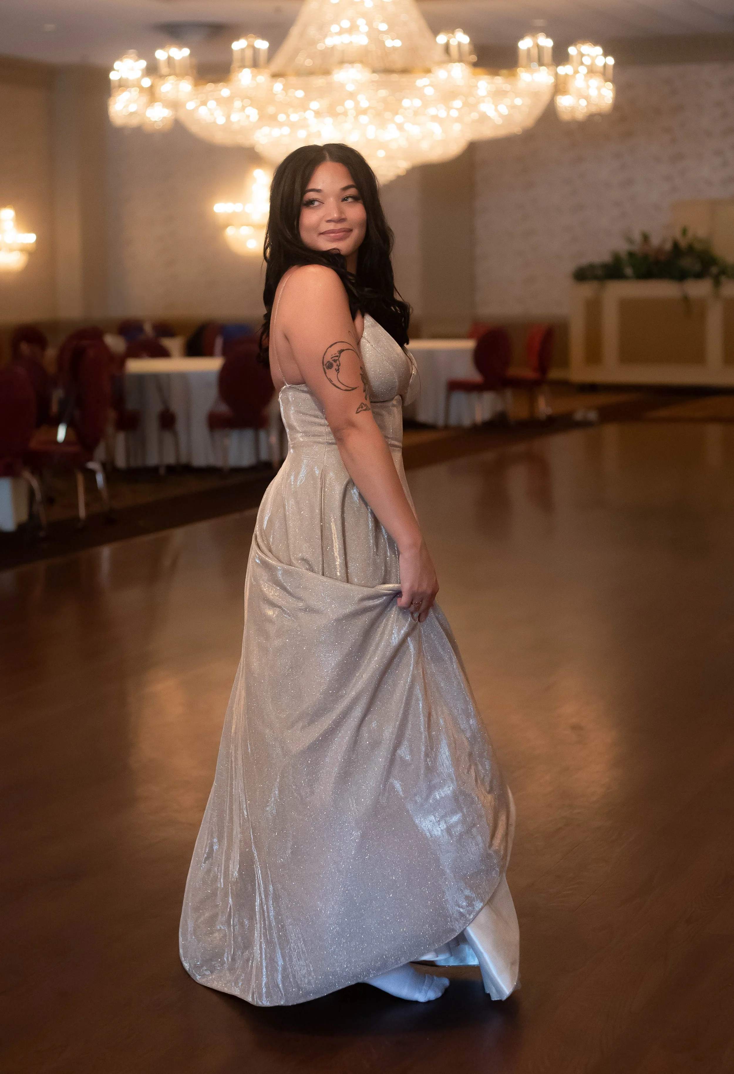 A woman with dark hair wearing a shimmery silver gown standing in a ballroom with a large chandelier overhead.