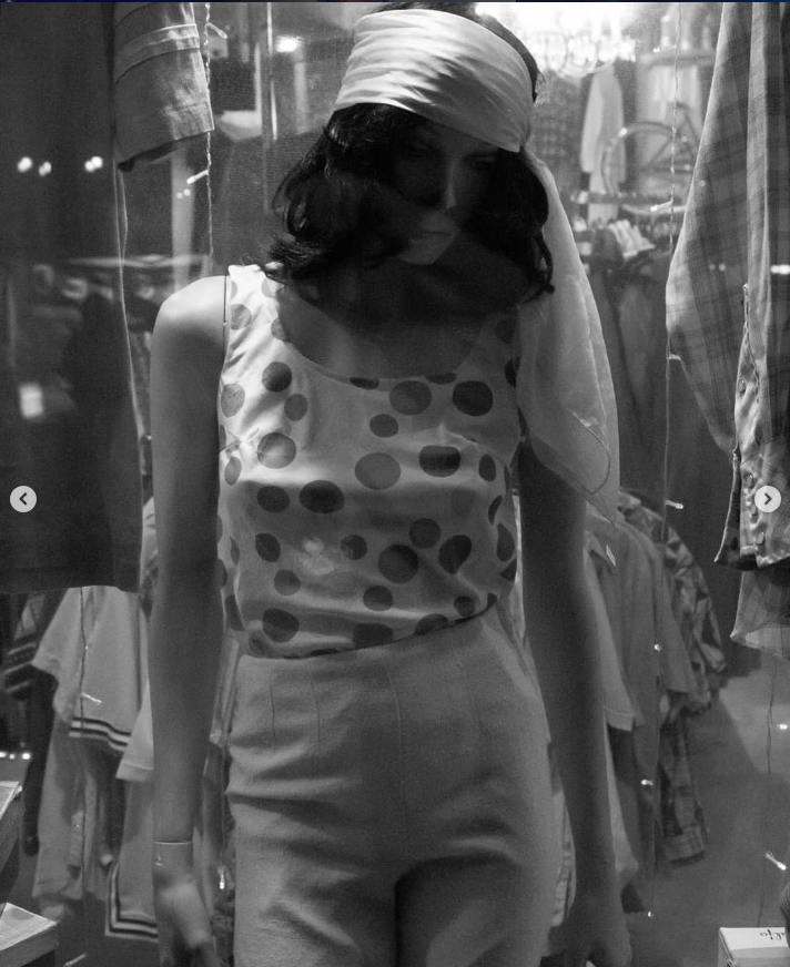 A mannequin wearing a polka dot sleeveless top and high-waisted pants inside a clothing store, with other clothes hanging around.