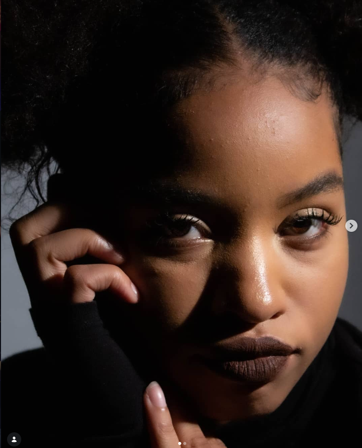 Close-up portrait of a young woman with natural hair, dark lipstick, and makeup, touching her face with her hand, with a serious expression and soft lighting highlighting her features.photo by Annelise Mirabal