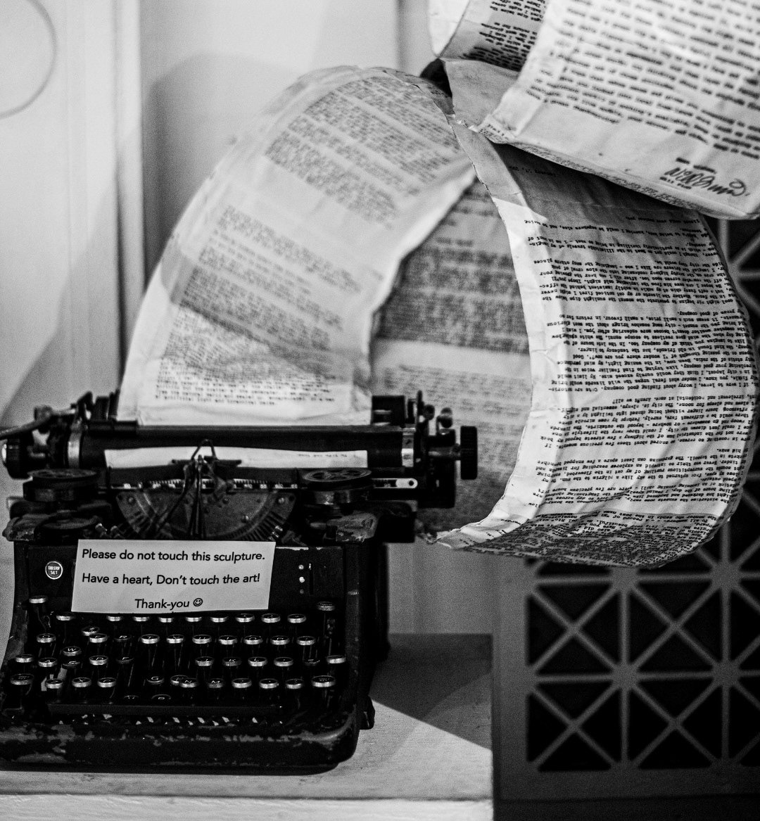 Old typewriter with a sculpture made of rolled newspaper strips around it and a sign that reads, 'Please do not touch this sculpture. Have a heart, Don't touch the art! Thank-you'.