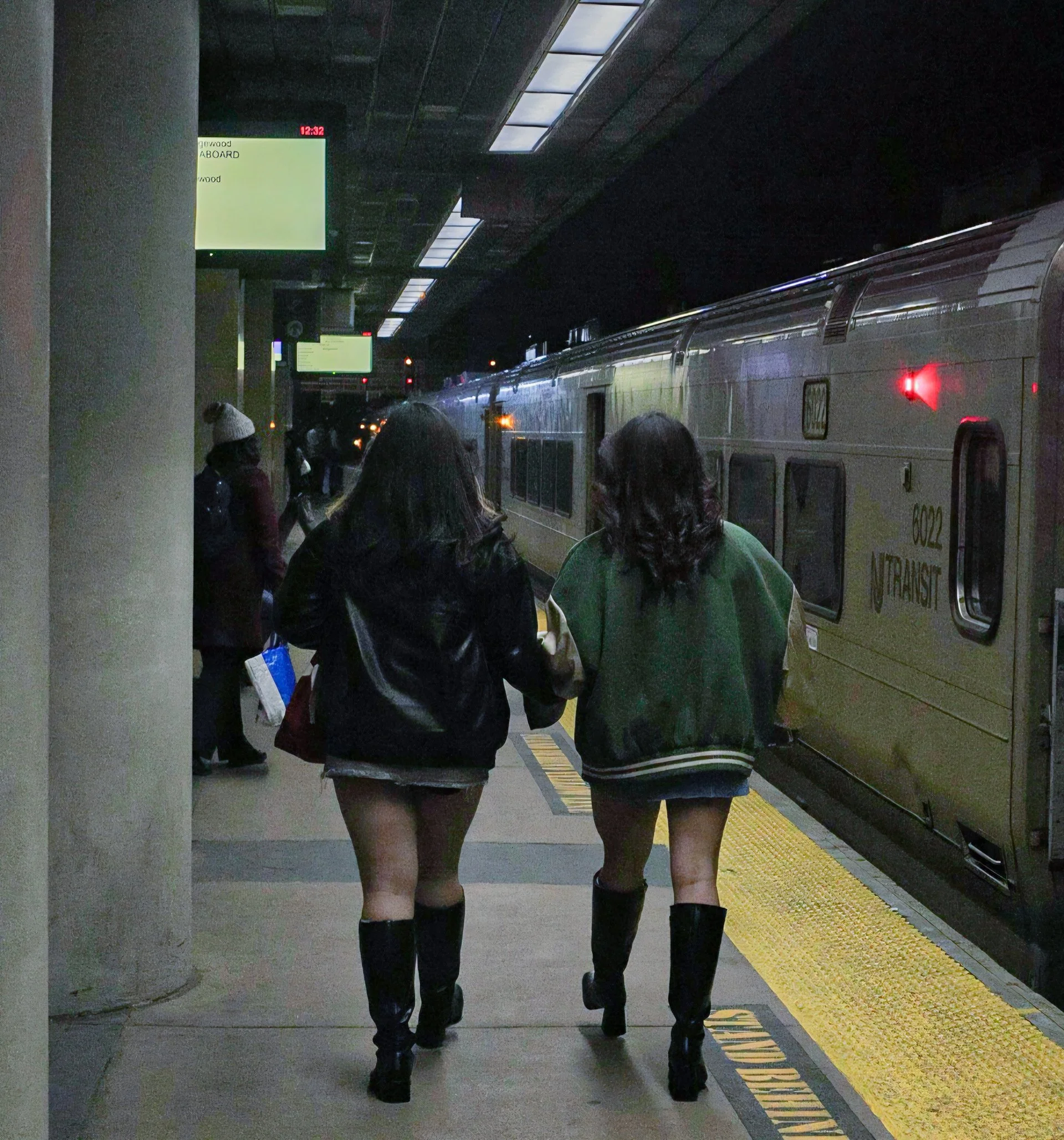 People walking on a subway platform with a silver train and illuminated signs in the background.