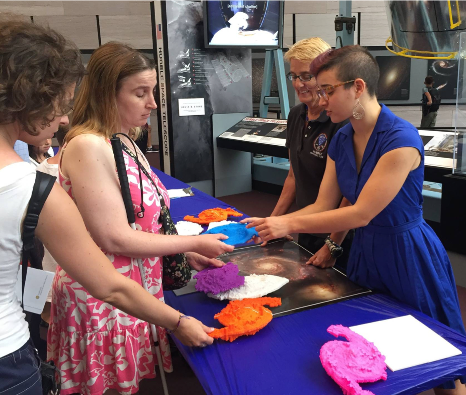 An individual from SciAccess stands behind a table filled with pictures and 3D models of spiral galaxies. They are pointing out features on a blue 3D model to a woman with a white cane who is feeling the features of the 3D model.