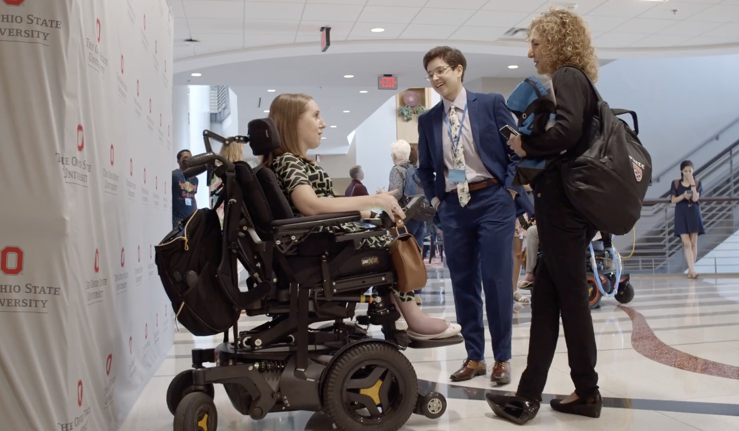 SciAccess Executive Director Anna Voelker, a nonbinary person in a blue suit, speaking with Miss Wheelchair USA 2018 Heather Tomko, a white woman in an electric wheelchair, and Anousheh Ansari, an Iranian-American engineer and first Iranian astronaut