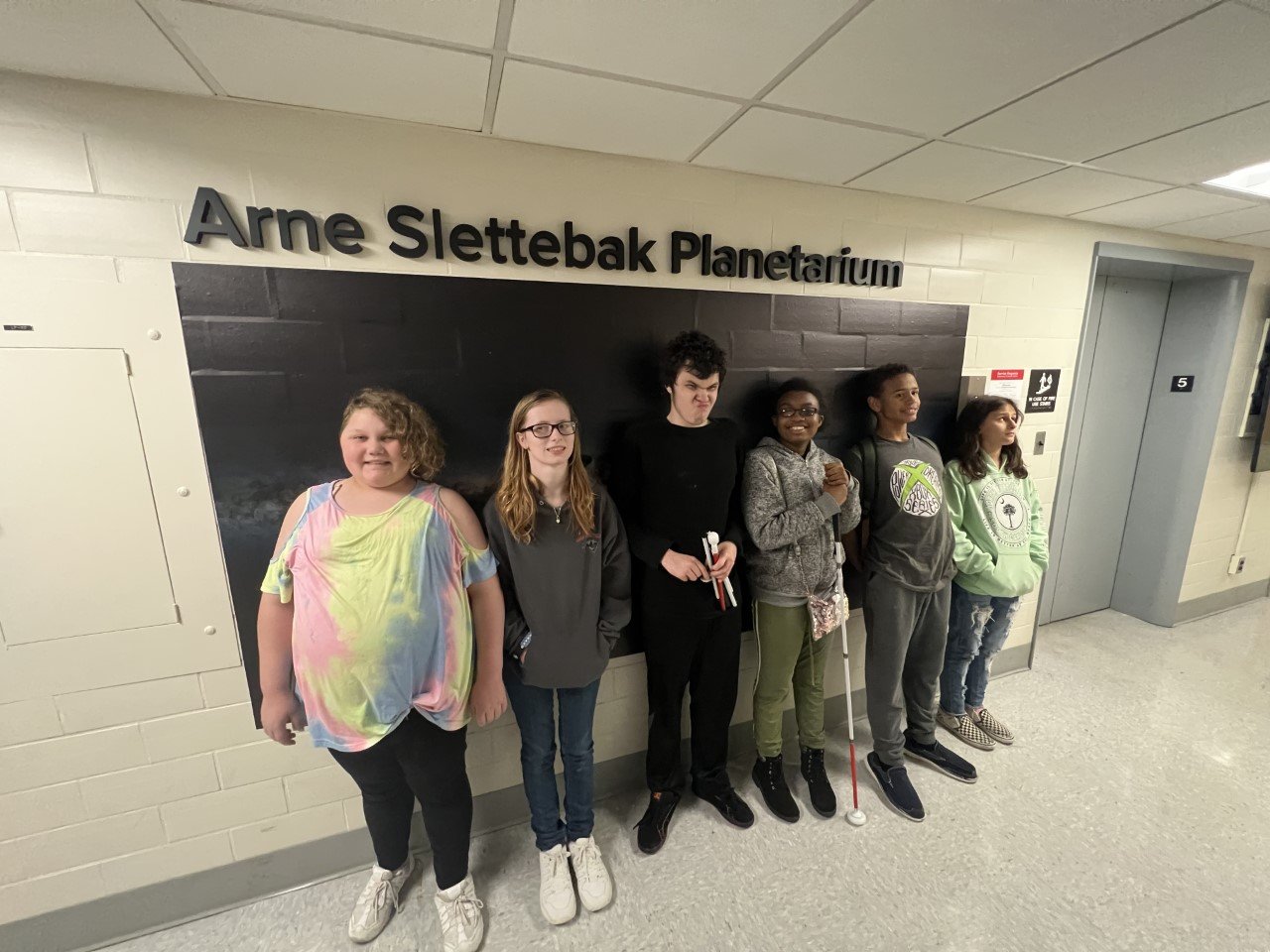 Young students holding white canes smile in front of a sign that says "Arne Slettebak Planetarium".
